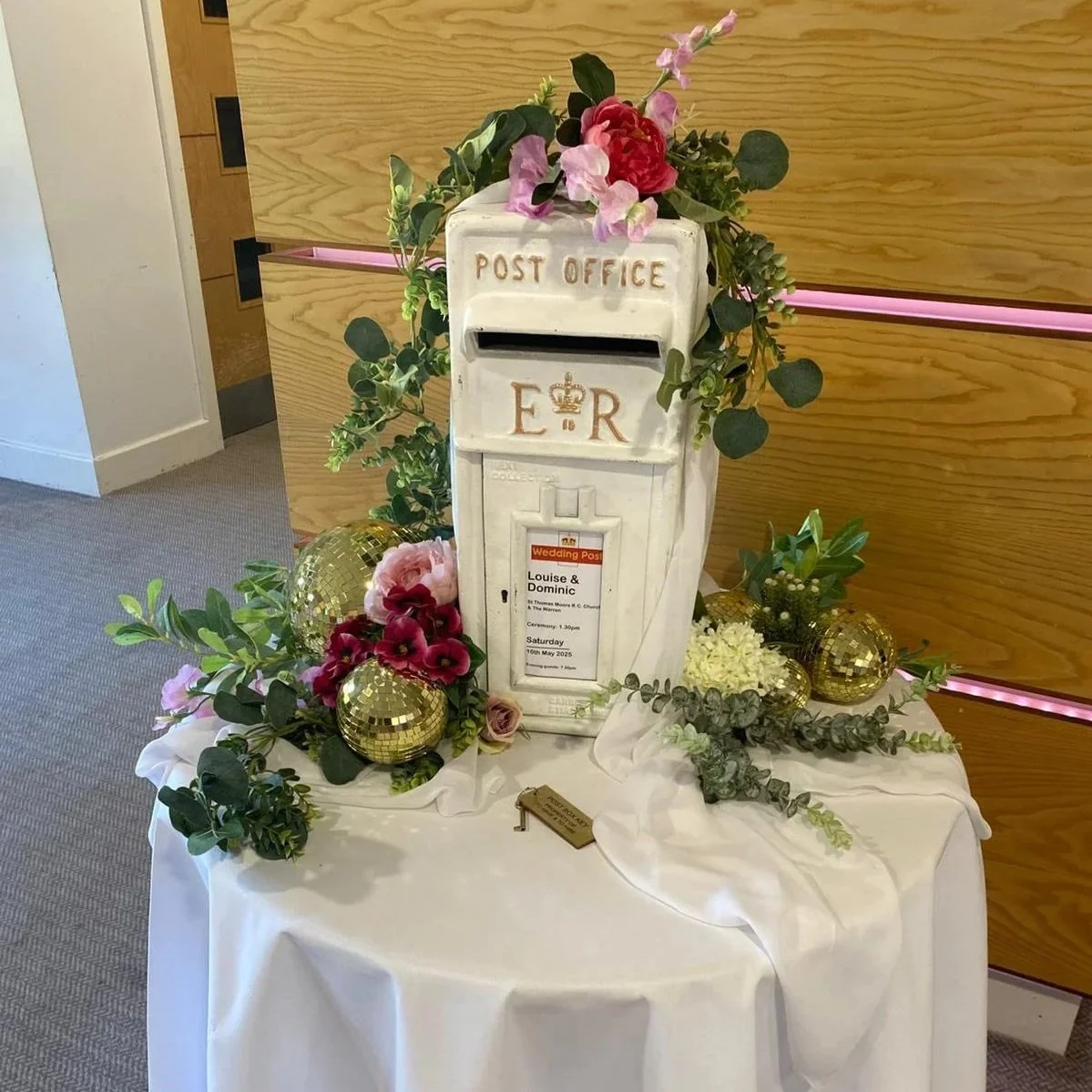 Decorative post box with floral arrangements, gold disco balls, and wedding information card, set on a white tablecloth at a wedding.