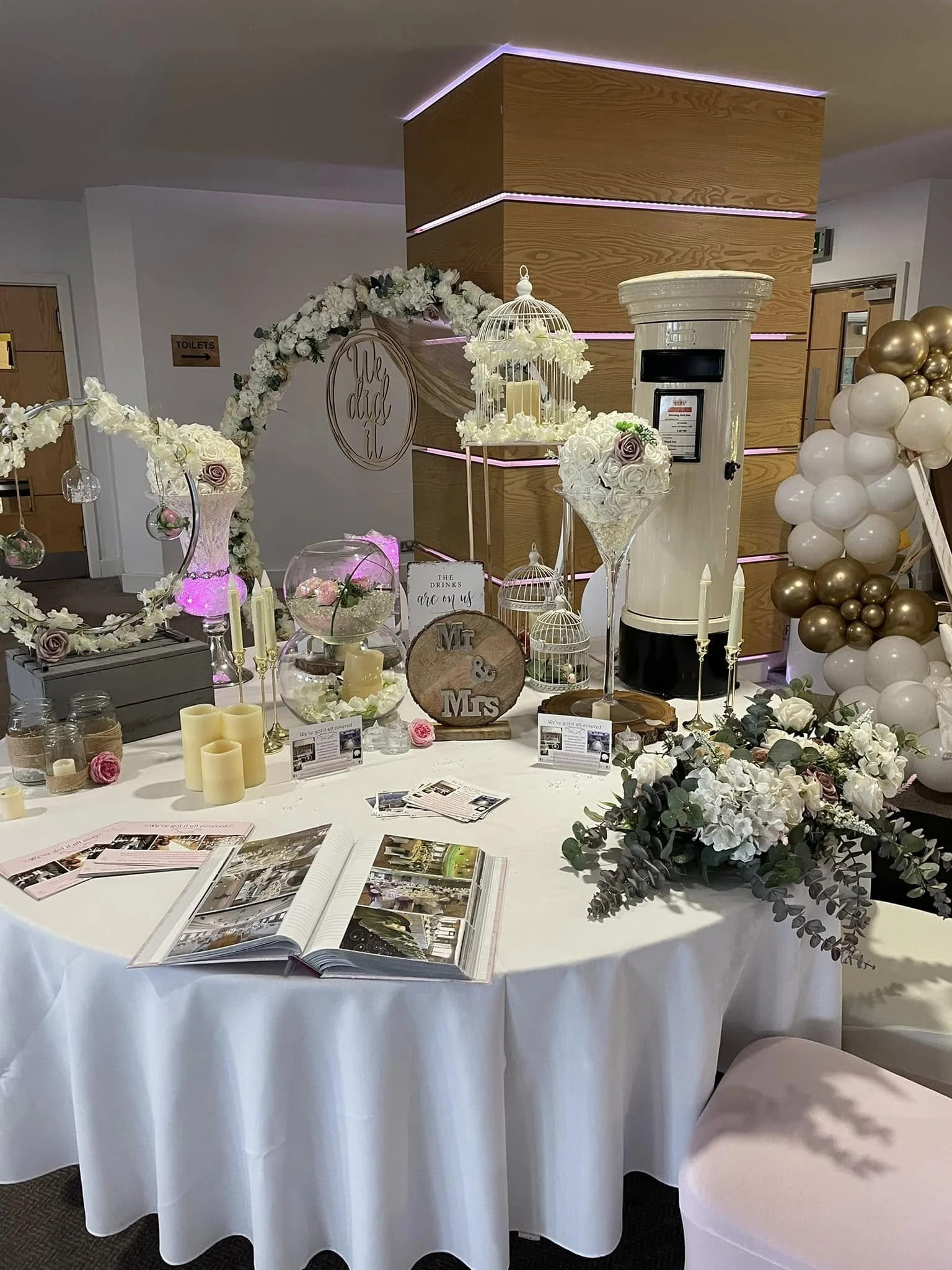 Decorative wedding table setup featuring floral arrangements, candles, birdcages, a photo album, and a white balloon arch in the background.