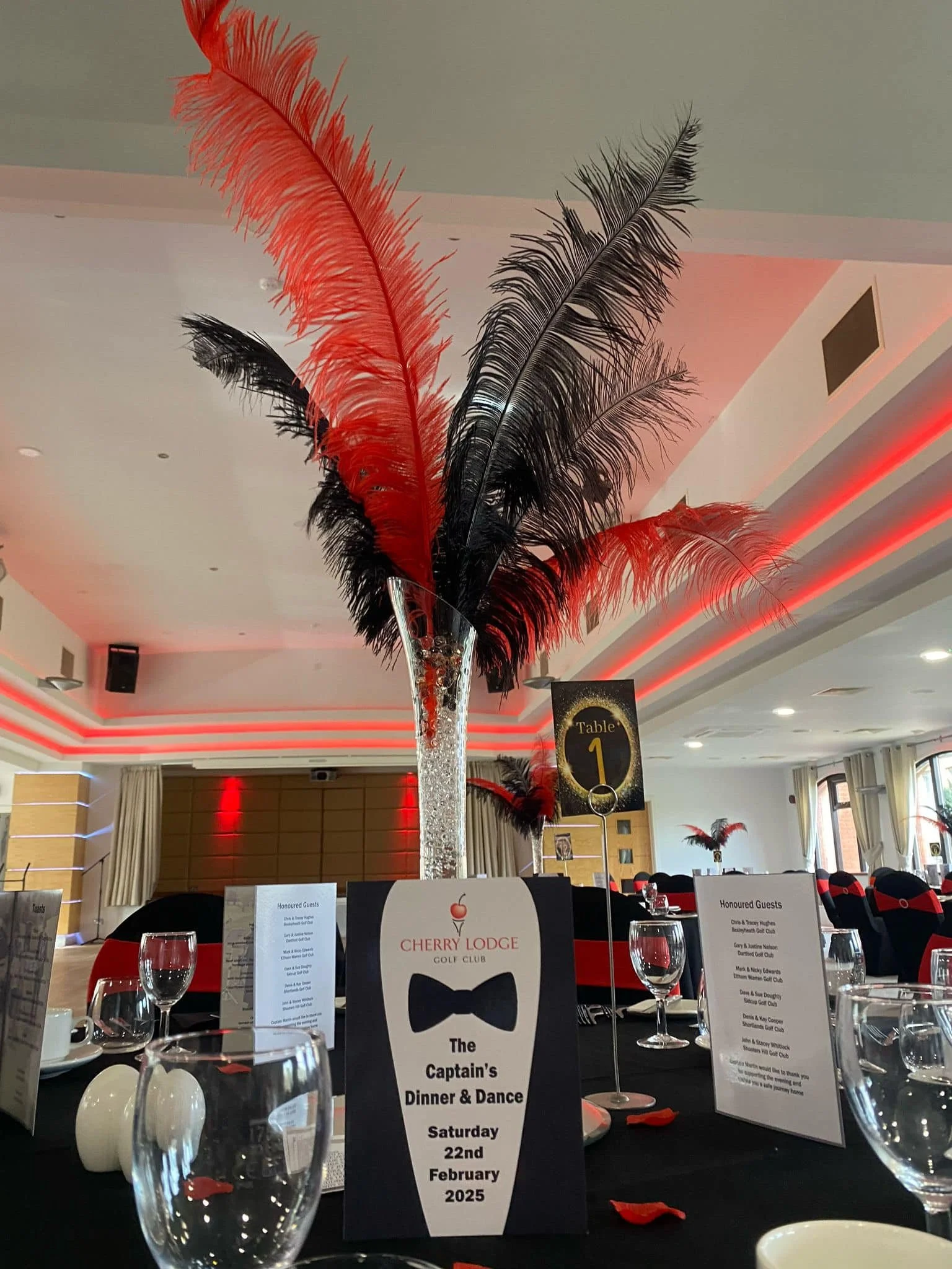 Decorated banquet hall with a tall centerpiece of red and black feathers in a crystal vase, set for a dinner event with a sign indicating 'Captain's Dinner & Dance' on February 22, 2025.