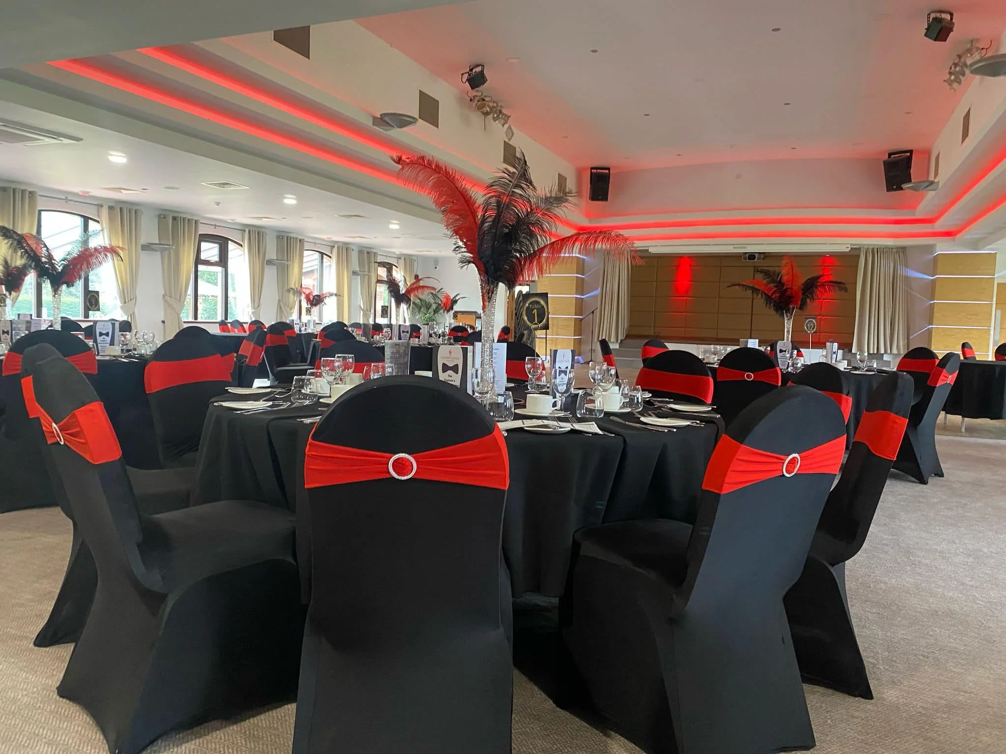 Banquet hall decorated for a formal event with black tables and chairs, red and black chair sashes, tall centerpieces with red and black feathers, and red lighting accents.