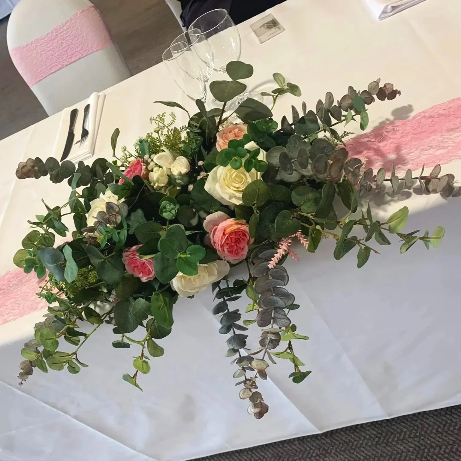 A floral centerpiece with white and pink roses, eucalyptus leaves, and other greenery on a table with white tablecloth and pink table runner, set for a formal event.