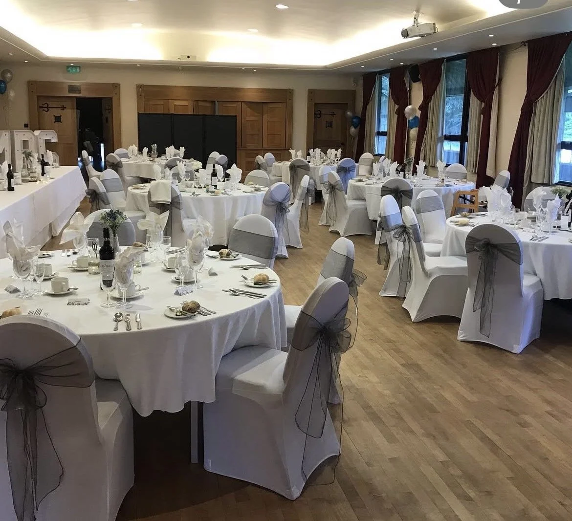 A banquet hall decorated for a formal event with round tables covered in white tablecloths, each surrounded by chairs with white covers and gray sashes, set with plates, glassware, and napkins folded in a fan shape, and balloons and balloons with ribbons decorating the room.