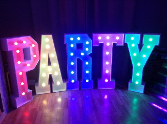 Large illuminated letters spelling out 'PARTY' in pink, yellow, blue, purple, and green lights, on a wooden floor with dark curtains in the background.