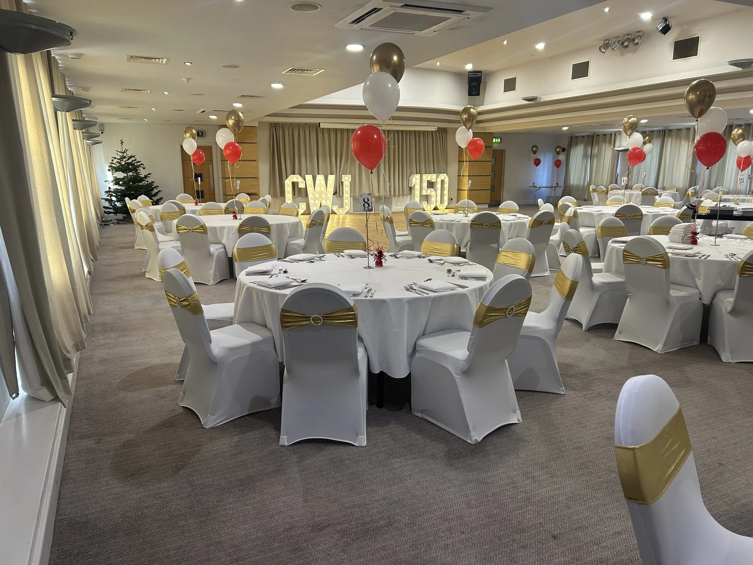A banquet hall decorated with white tablecloths, gold and white chair covers, and balloons in red, gold, and white. There is a decorated Christmas tree in the corner and illuminated signs reading 50 and CWI in the background.
