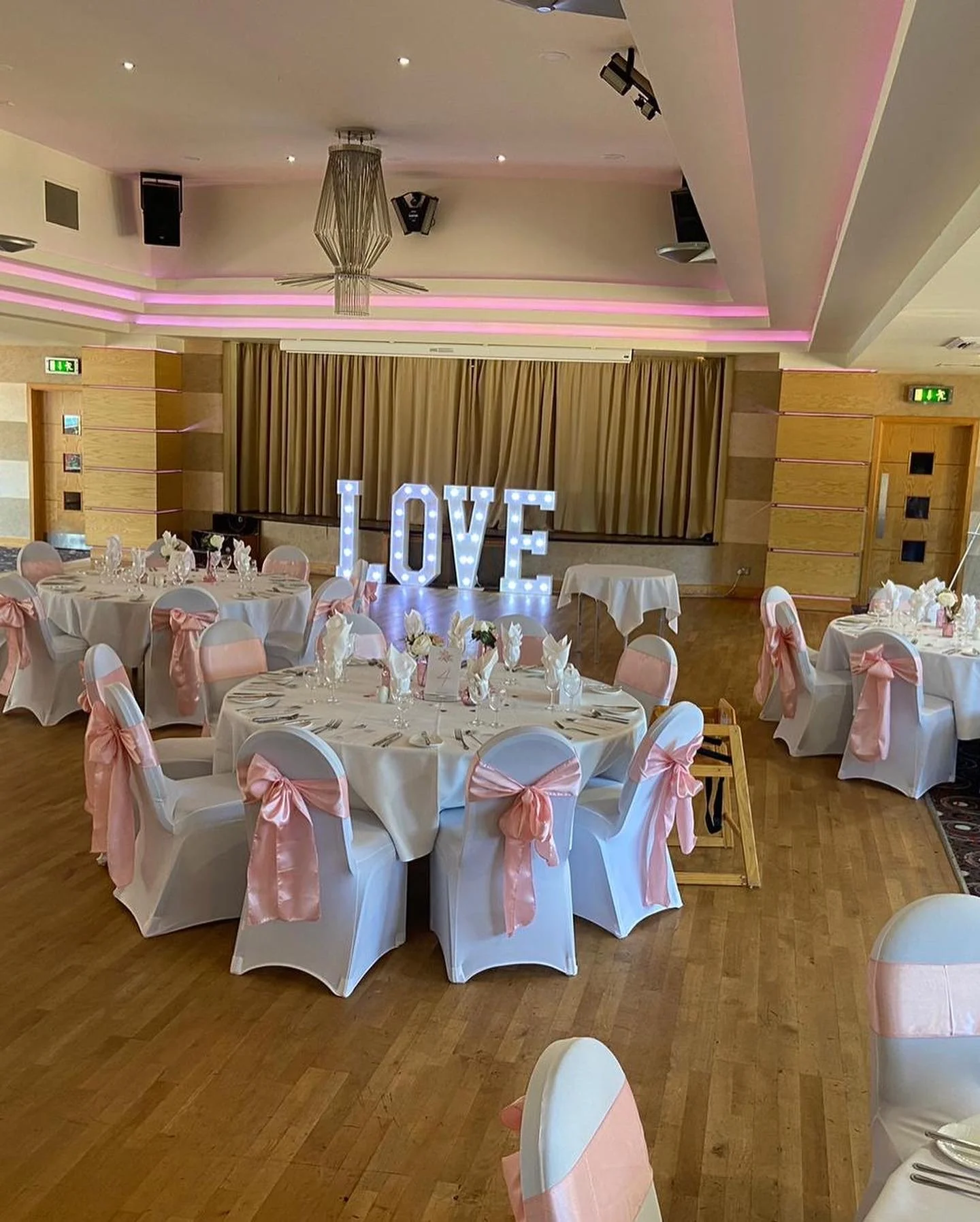 A decorated event hall with round tables covered in white tablecloths, adorned with pink bows on white chairs. Large illuminated letters spell out 'LOVE' on stage, with a gold curtain backdrop and pink lighting accents.