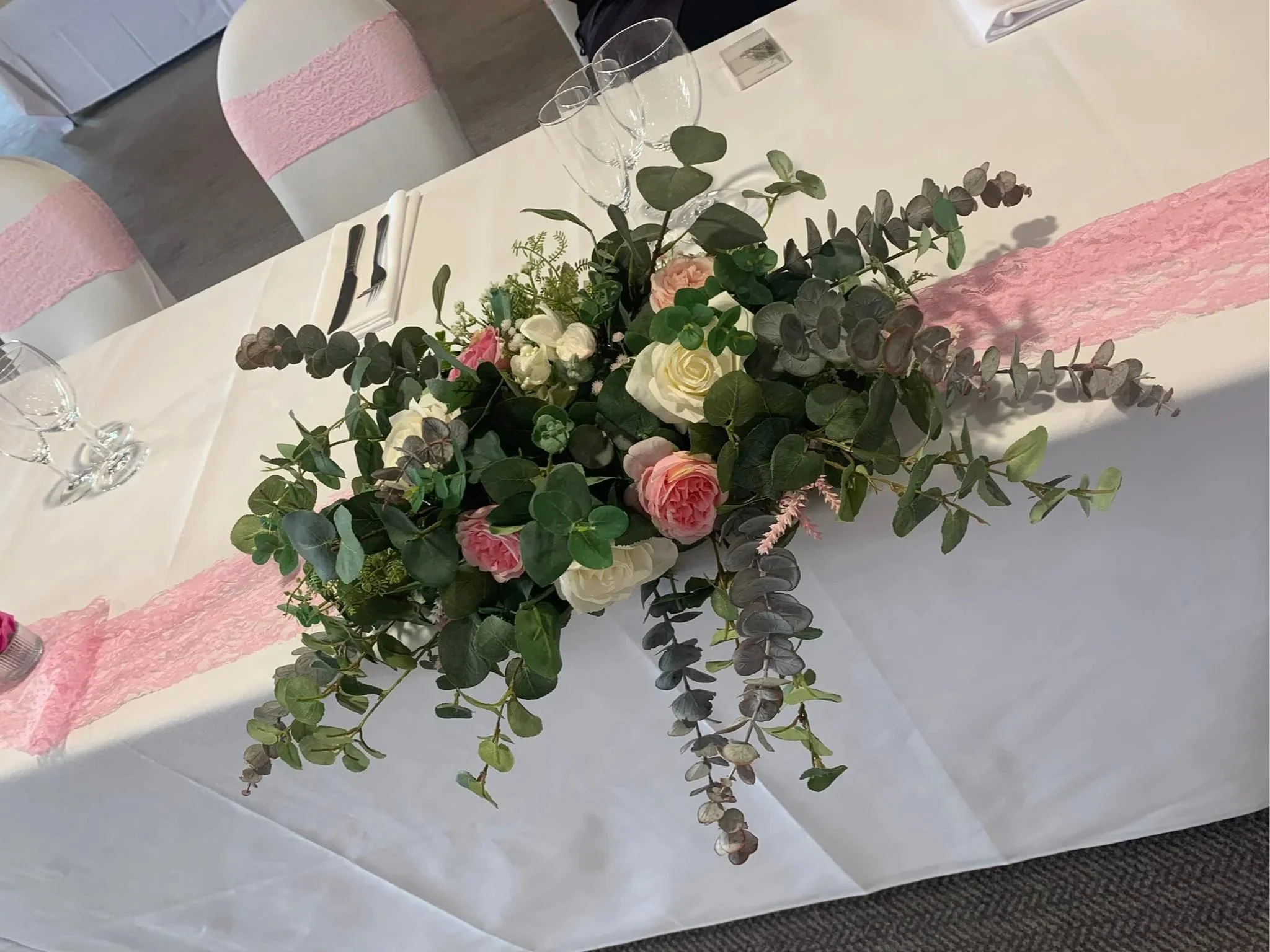 Elegant table centerpiece with pink and white roses, green foliage, and eucalyptus, set on a white tablecloth with pink lace runners at a formal event.