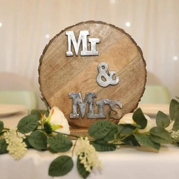 A round wooden wedding sign with metal letters spelling out "Mr & Mrs" placed on a table decorated with white roses and greenery.