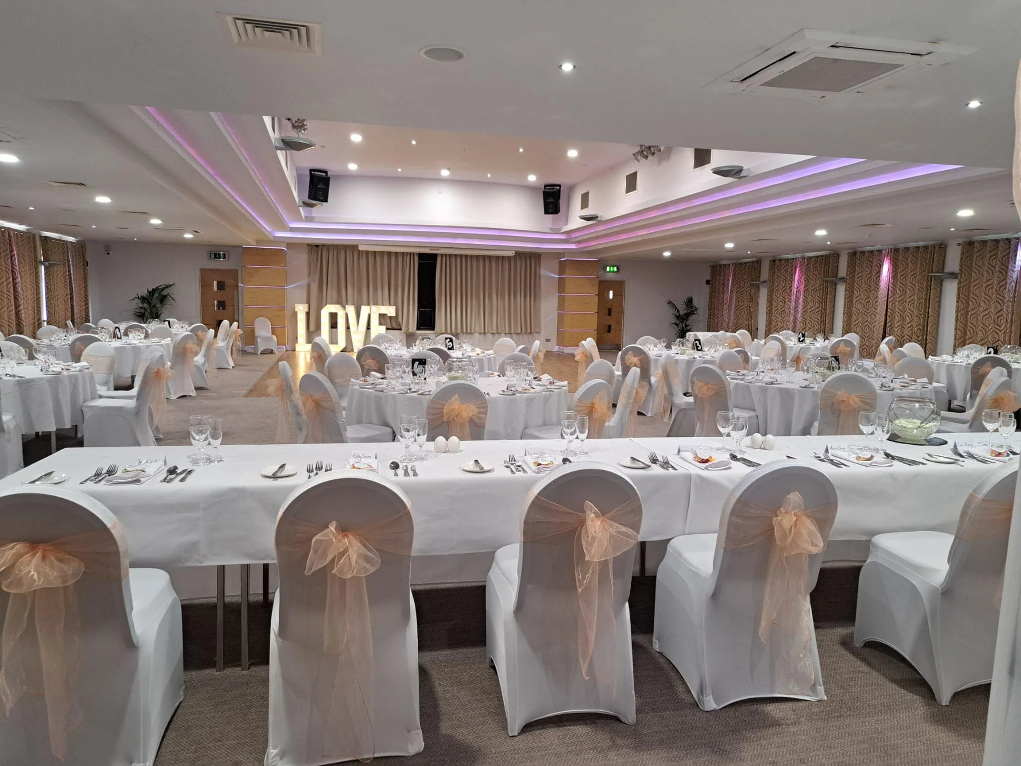 Elegant event hall decorated with white tablecloths, chairs with peach-colored bows, and illuminated "LOVE" sign in the background.