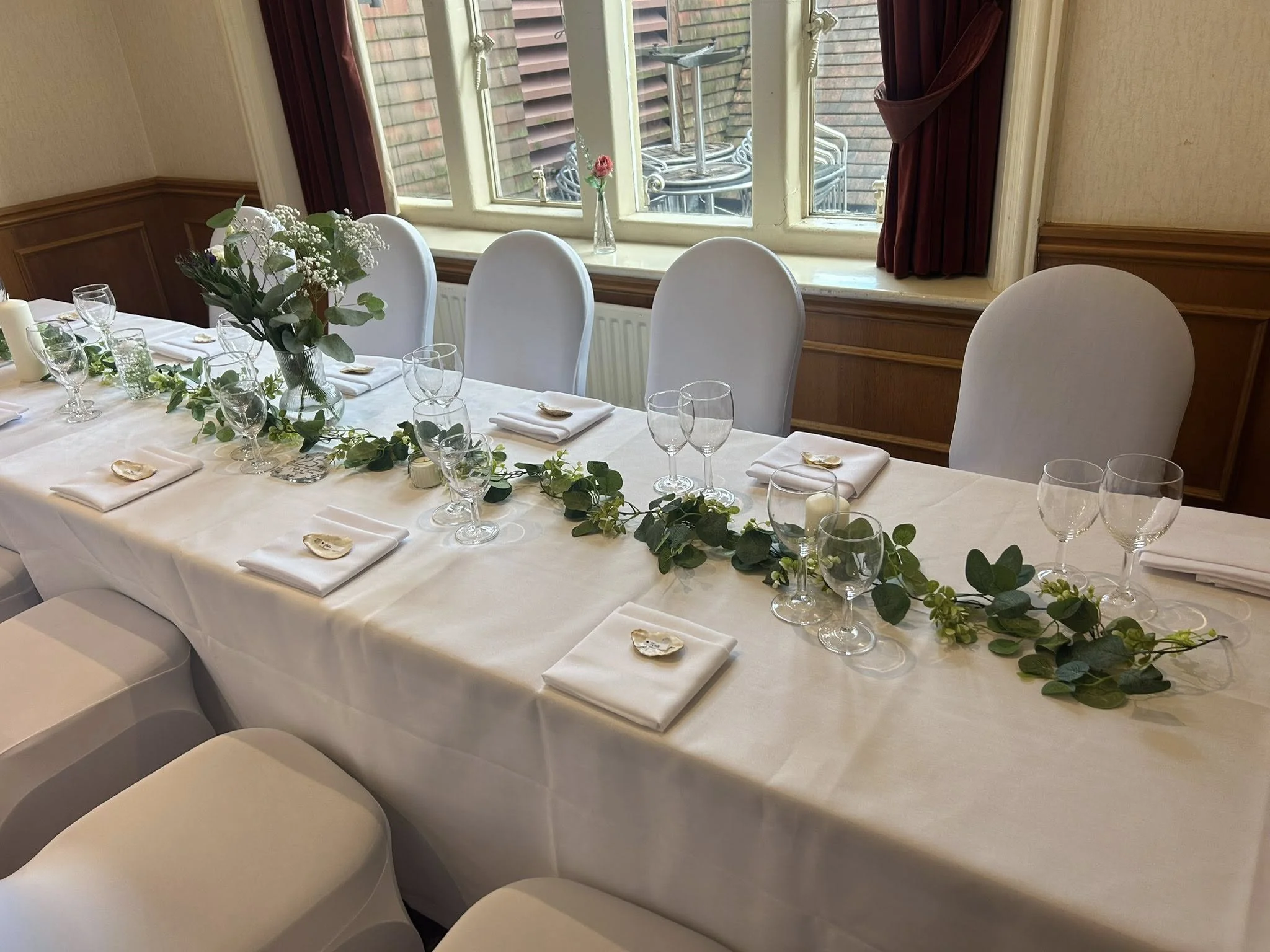 A elegantly set dining table with white tablecloth, glassware, folded white napkins, and a centerpiece of flowers and greenery, near a window with a view outside.