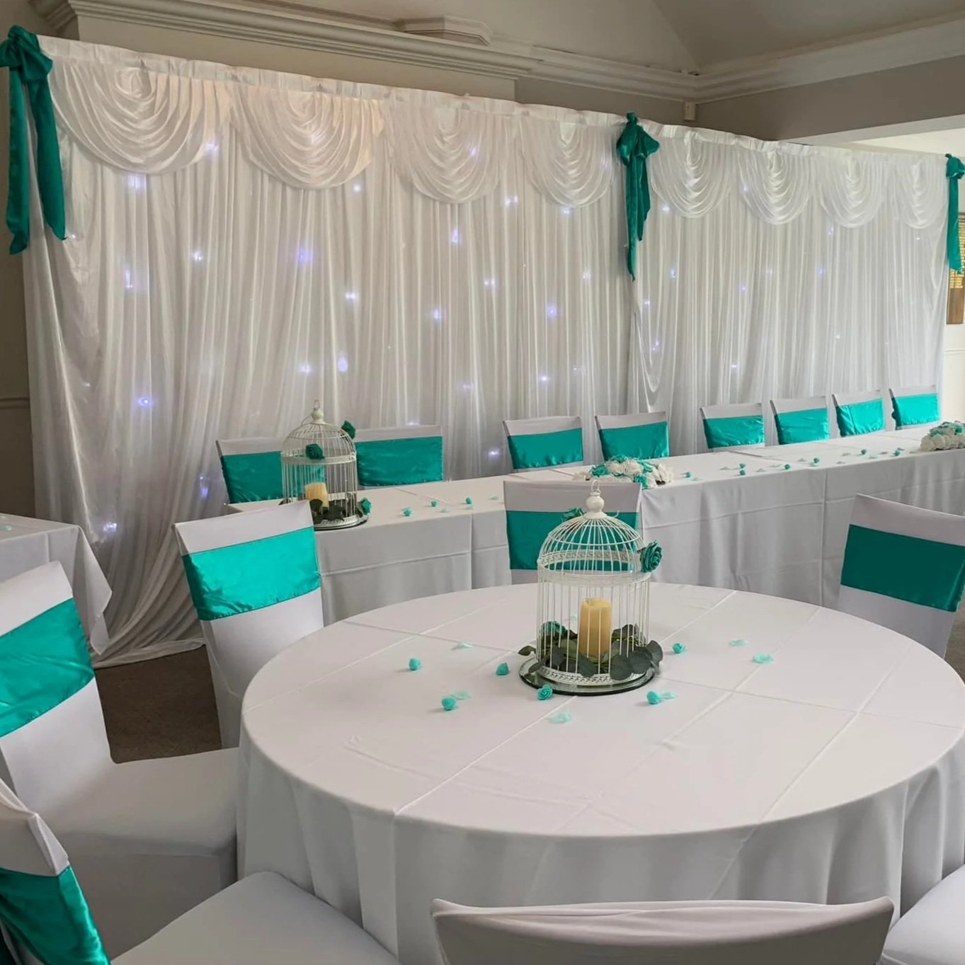 Elegant banquet hall with white draped backdrop and teal accents, decorated for a celebration with round and rectangular tables, teal chair sashes, birdcage centerpieces, and string lights.