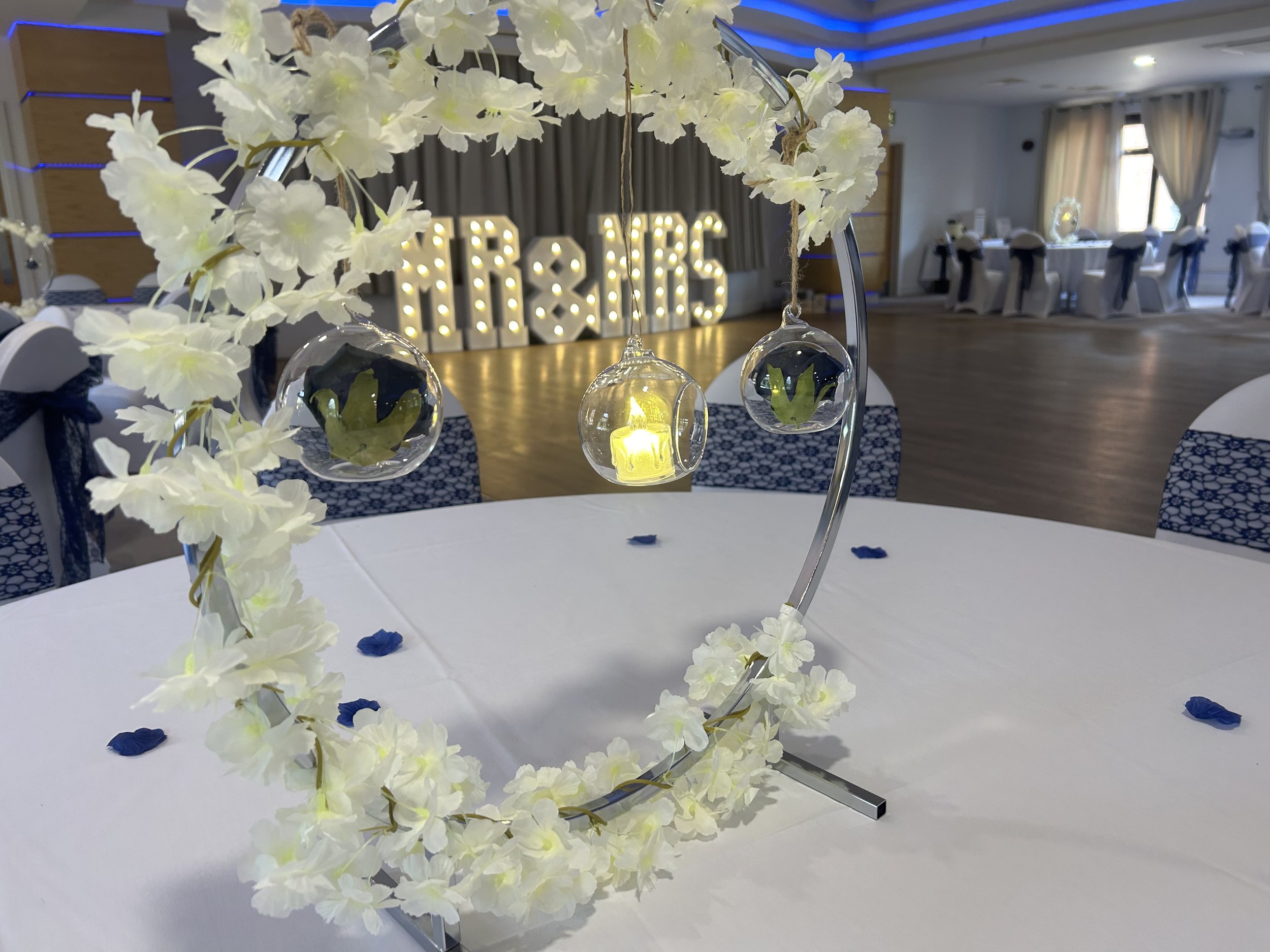 Wedding reception table centerpiece with white flowers, hanging glass globes with green plants, and a glowing candle in the middle. In the background, large illuminated letters spelling 'MR & MRS' are visible, along with draped curtains and arranged tables with chairs covered in white cloth and black sashes.