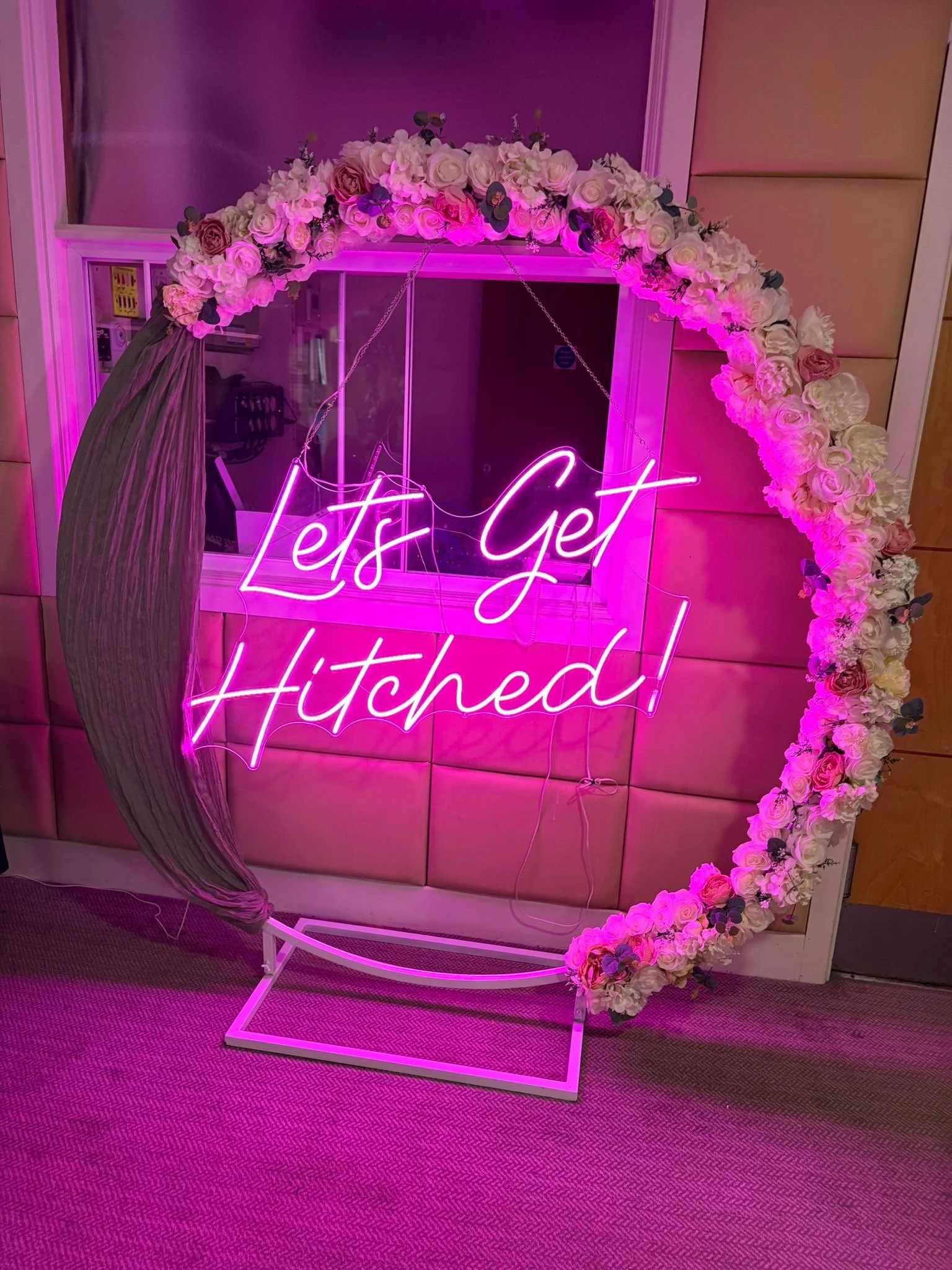A decorative display with a neon pink sign reading 'Let's Get Hitched!', a floral arch with pink and white roses, and a cloth draped on the left side.