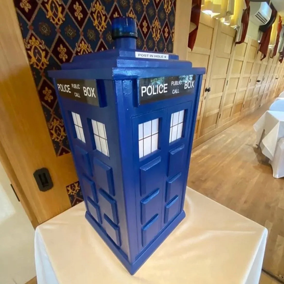 A decorative blue mailbox designed to look like the TARDIS from Doctor Who, placed on a white table inside a room with wooden flooring and cabinets, and patterned wallpaper in the background.
