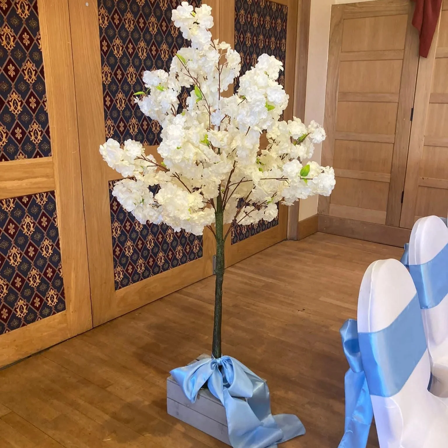 Artificial white flower tree with a gray base decorated with a blue bow, placed indoors on a wooden floor next to chairs with white covers and blue sashes.