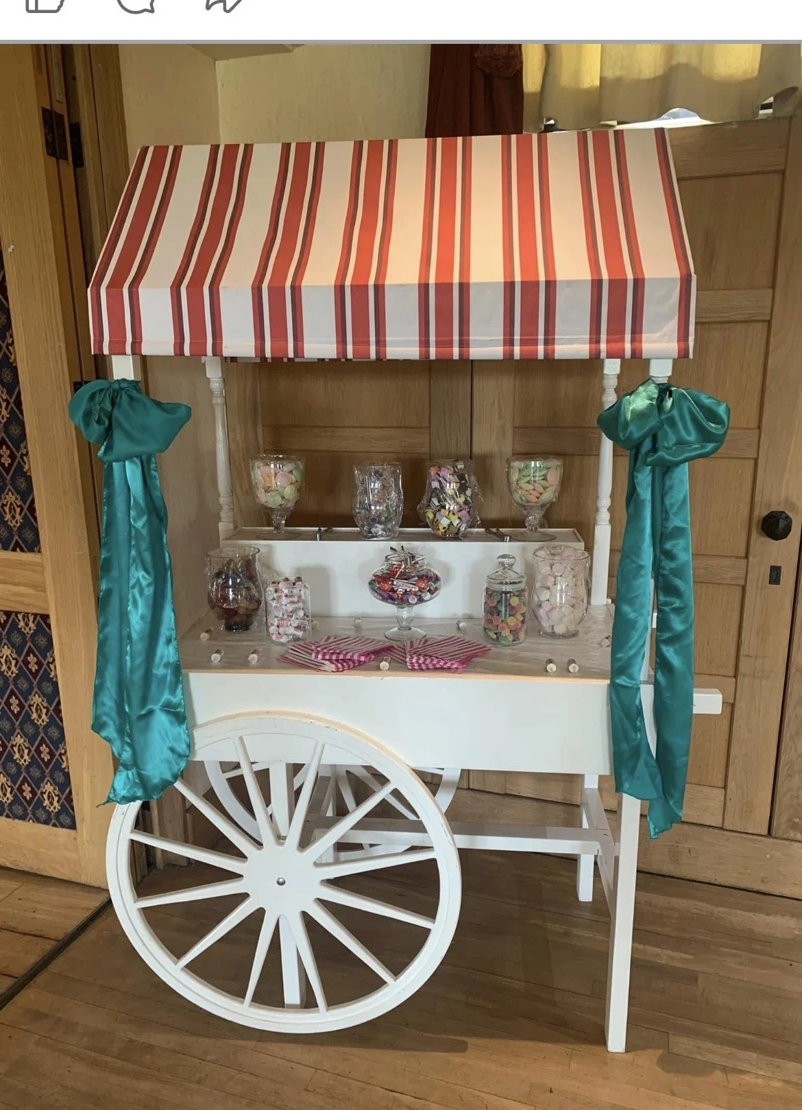 A white cart with one large wheel, decorated with teal satin ribbons on each side, and topped with a red and white striped canopy. The cart is filled with jars and bowls of colorful candies, set in a room with wood-paneled walls and flooring.