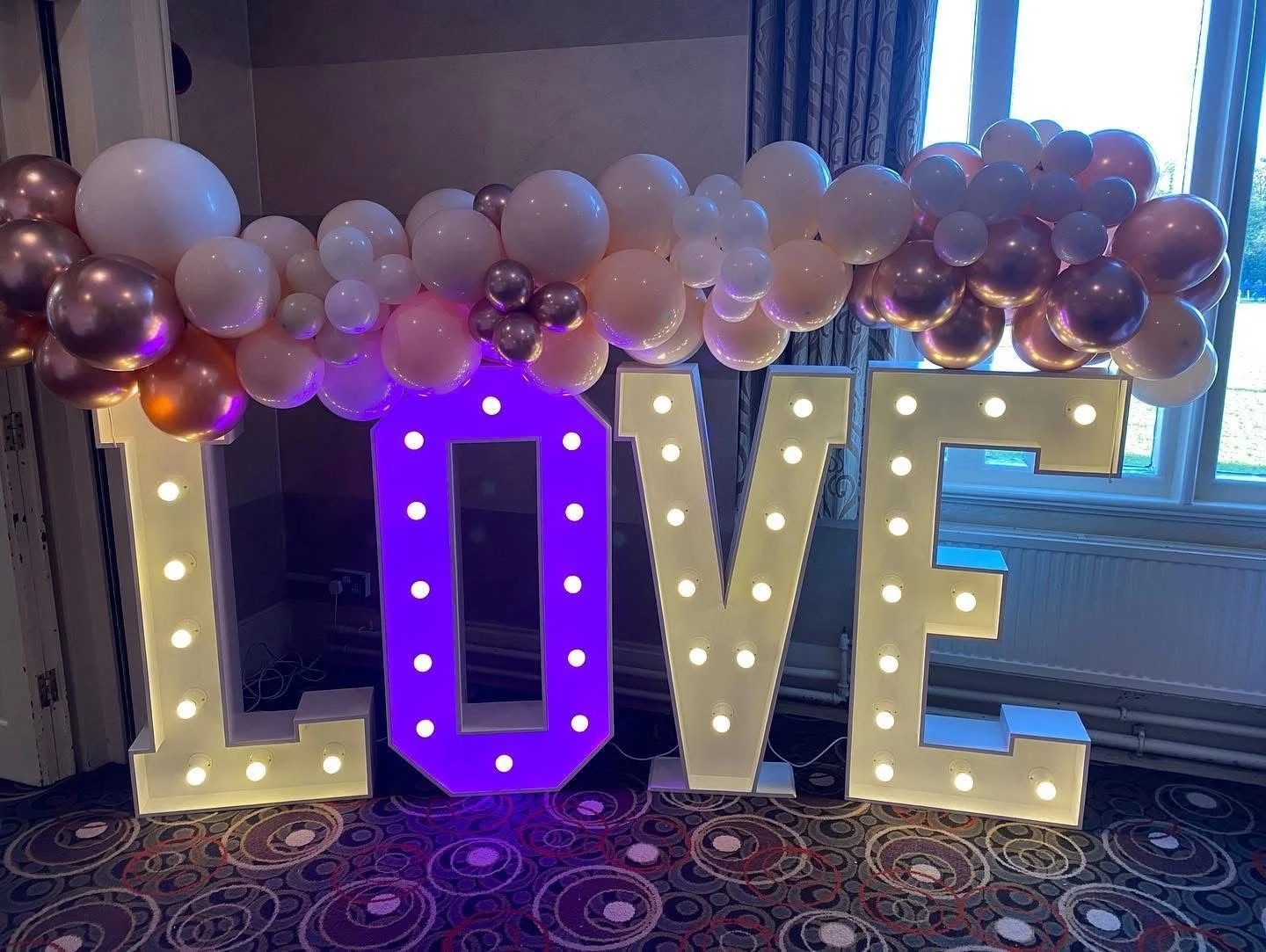 Decorative display with large illuminated letters spelling 'LOVE' and a cloud of pink, white, and rose gold balloons above them.