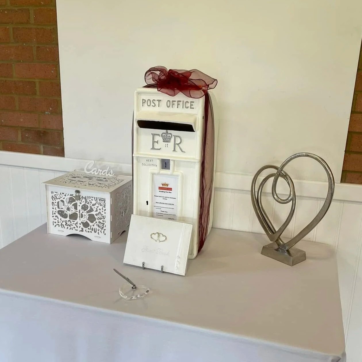 A vintage white post box with black mail slot and red ribbon on top, placed on a table with wedding-themed decorations including a white box with cut-out heart and flower design labeled 'Cards', a white wedding card with gold wedding rings illustration, and a metal sculpture of intertwined hearts.