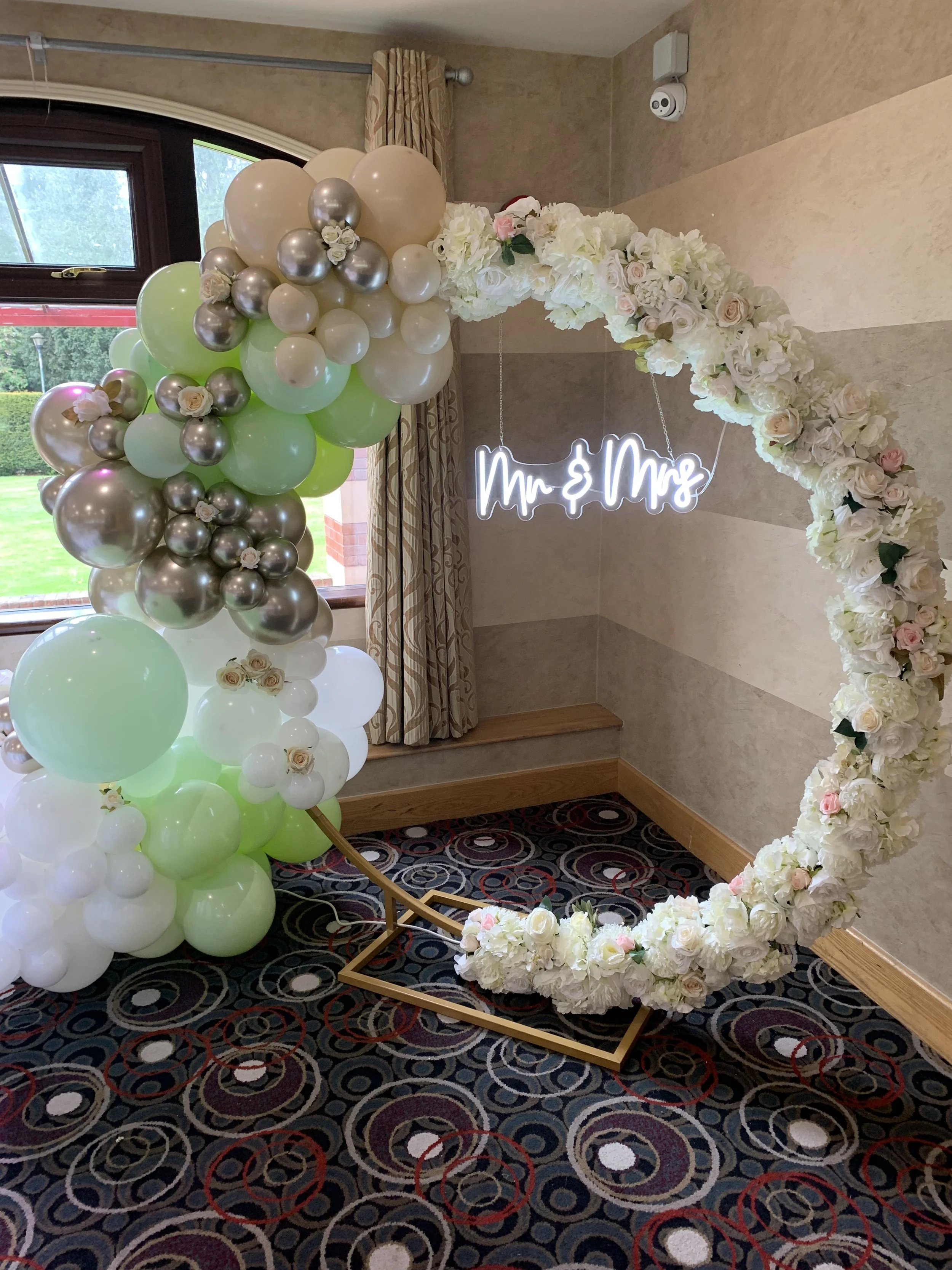 A decorated arch with white and pink flowers and a neon sign reading 'Mr & Mrs,' with a cluster of balloons in white, silver, light green, and chrome, standing near a window with curtains in a room.