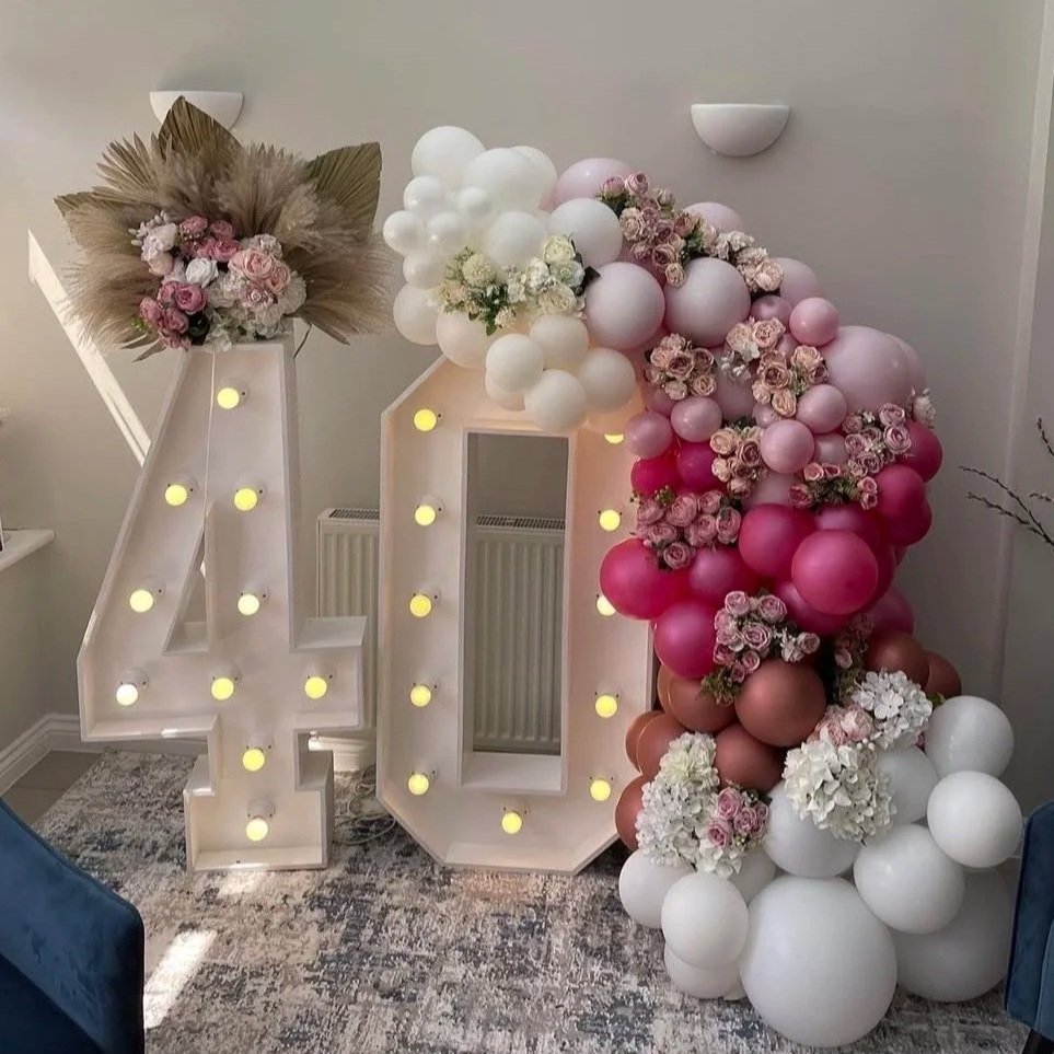 Decorative arrangement of large illuminated 4 and 0 letters with floral and balloon decorations in pink, white, and beige, displayed indoors.