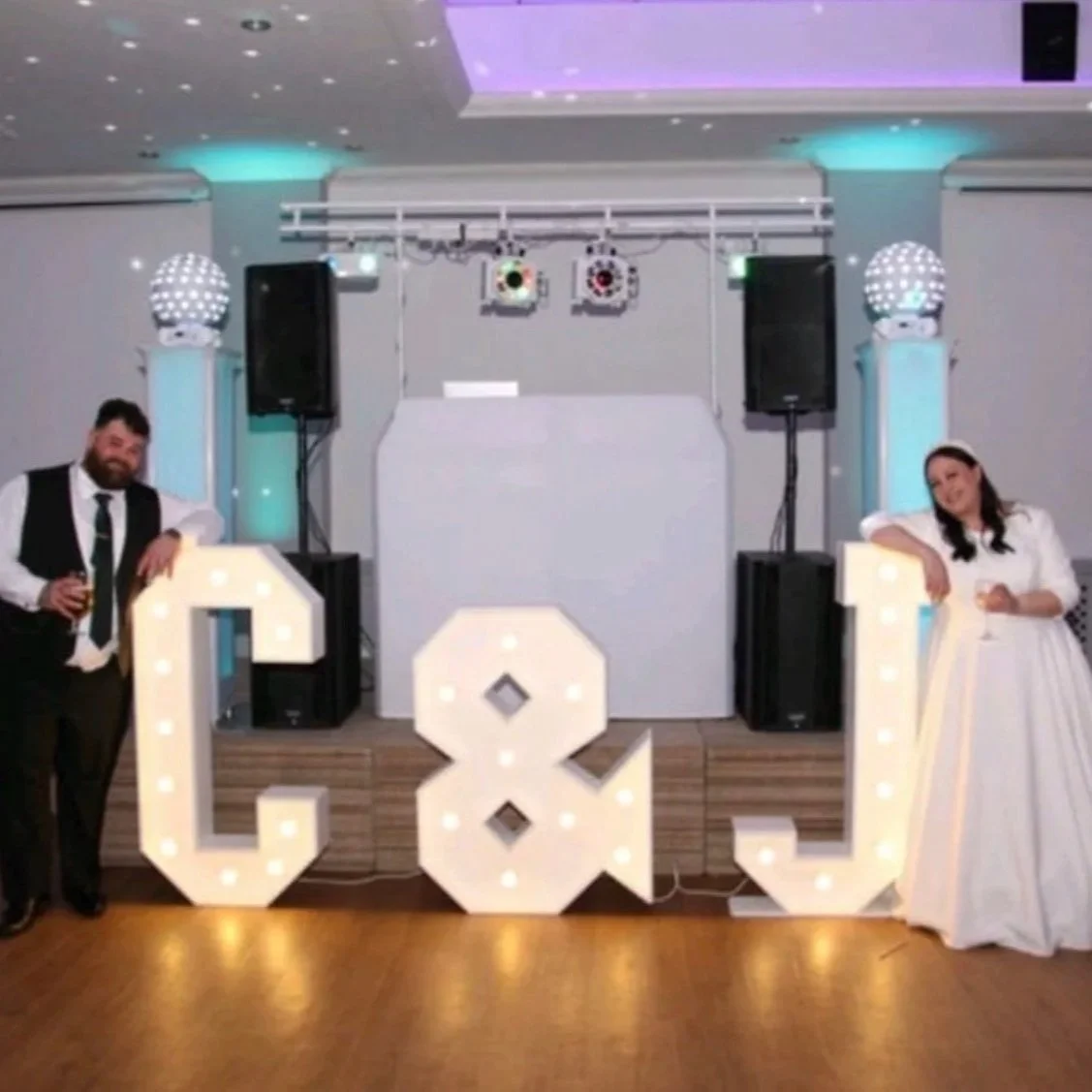A wedding reception with a DJ setup in the background, large illuminated letters 'C' and 'Q' in front, and a man and woman dressed in formal attire posing and holding drinks.