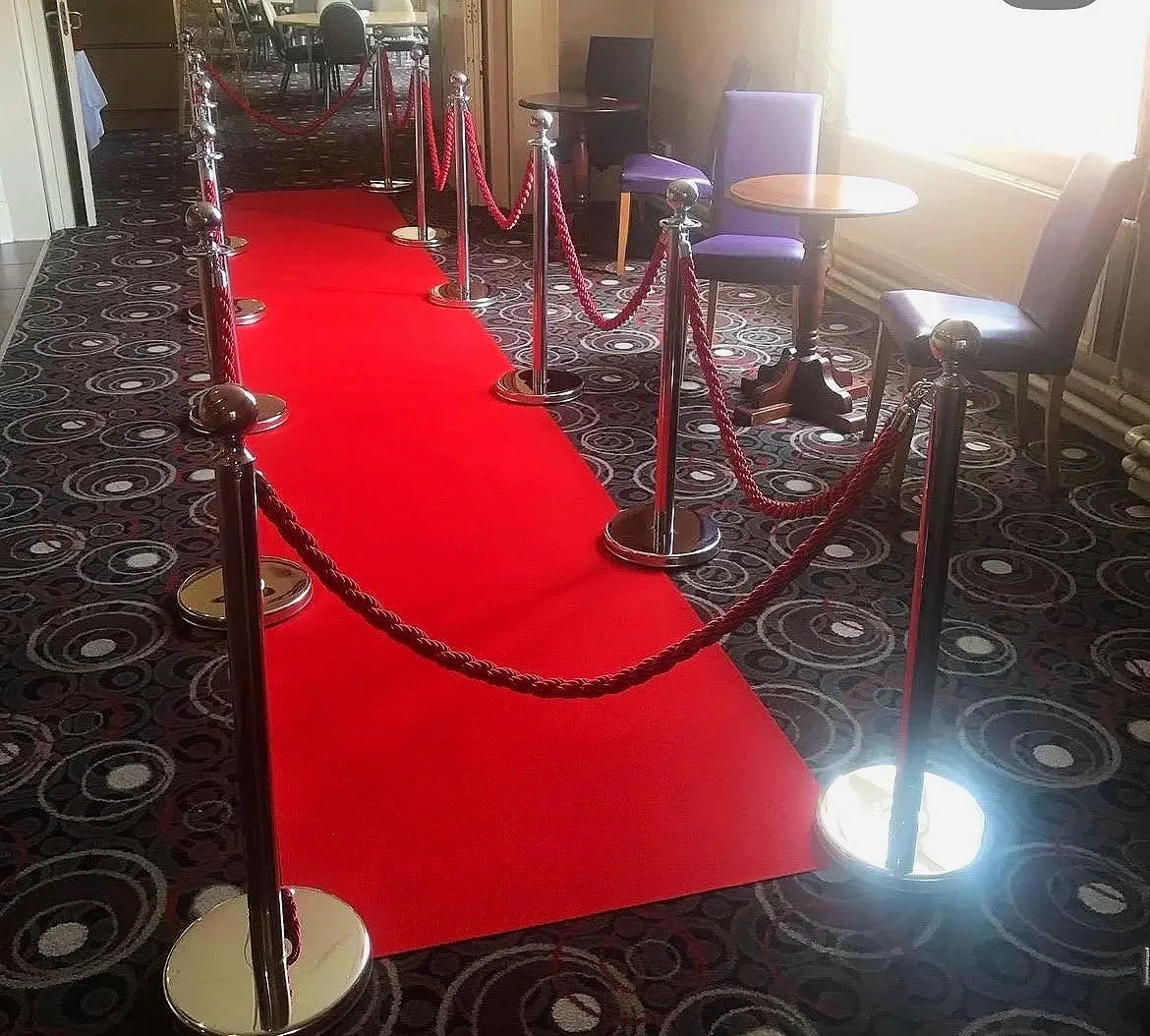 Red carpeted pathway with stanchions and velvet ropes, leading to a seating area with chairs and small tables near a window with sunlight.