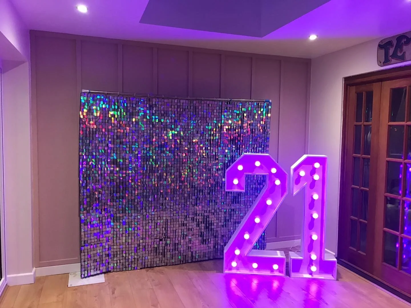Decorative scene with a colorful, shimmering mosaic backdrop and a large illuminated number '21' sign in a room with wooden flooring and sliding glass doors.