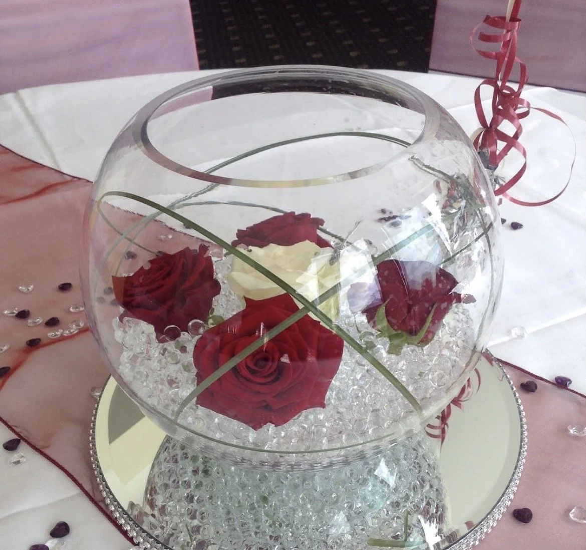A decorative glass bowl filled with white and red roses surrounded by small crystal beads, placed on a table with a pink and white tablecloth and surrounded by pink curled ribbons and scattered beads.