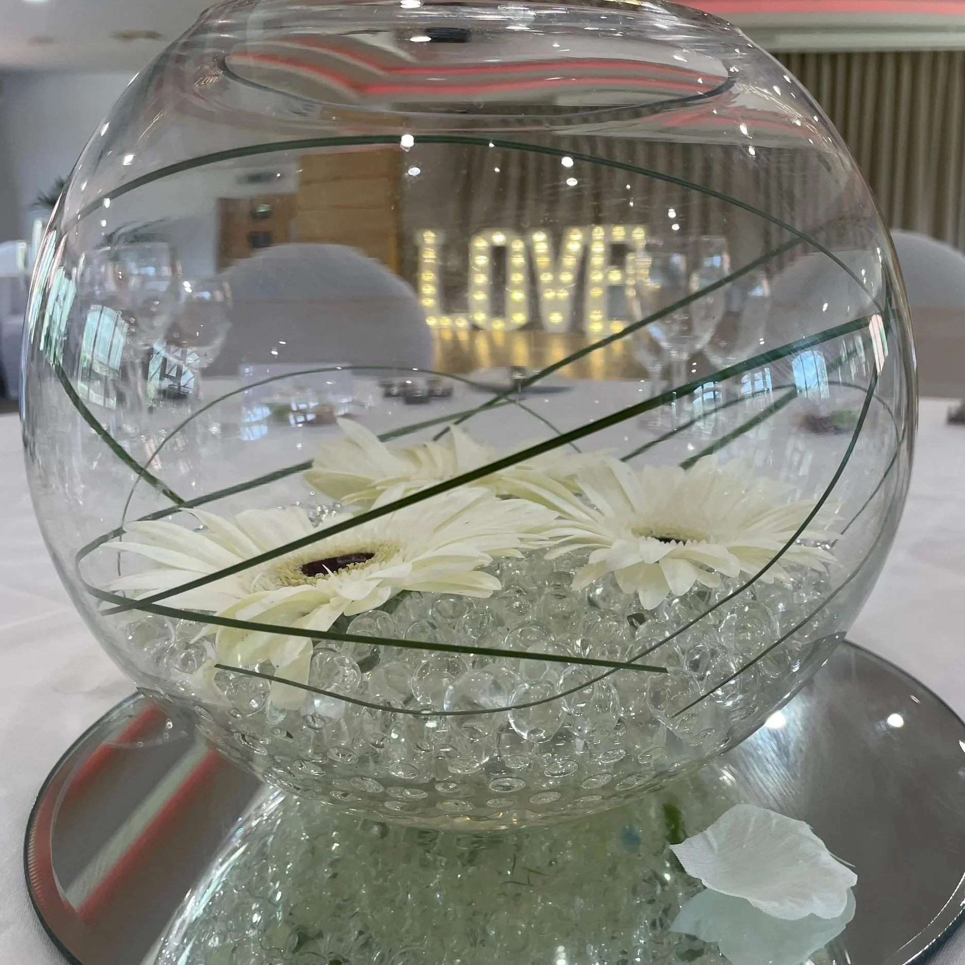 A glass bowl filled with water, white flowers, and decorative glass beads, on a table with a mirror underneath, with illuminated word 'LOVE' in the background.