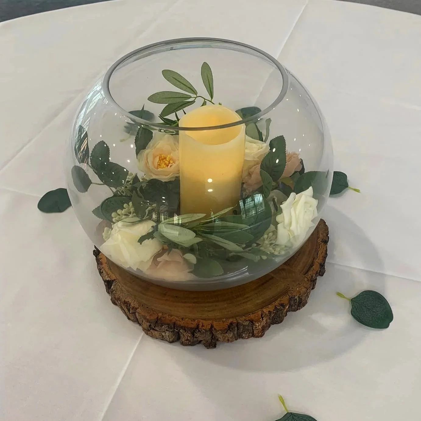 Glass bowl with a candle and floral arrangement on a wooden slab, placed on a white table.