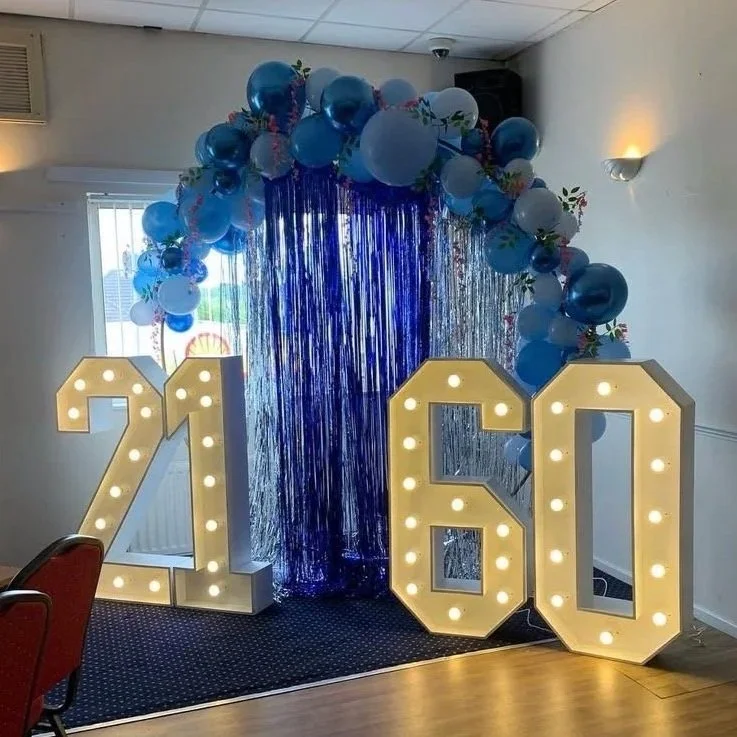 Large illuminated numbers 26 with balloons and metallic streamers in blue, white, and floral patterns, set up as a decoration for a 26th birthday celebration.