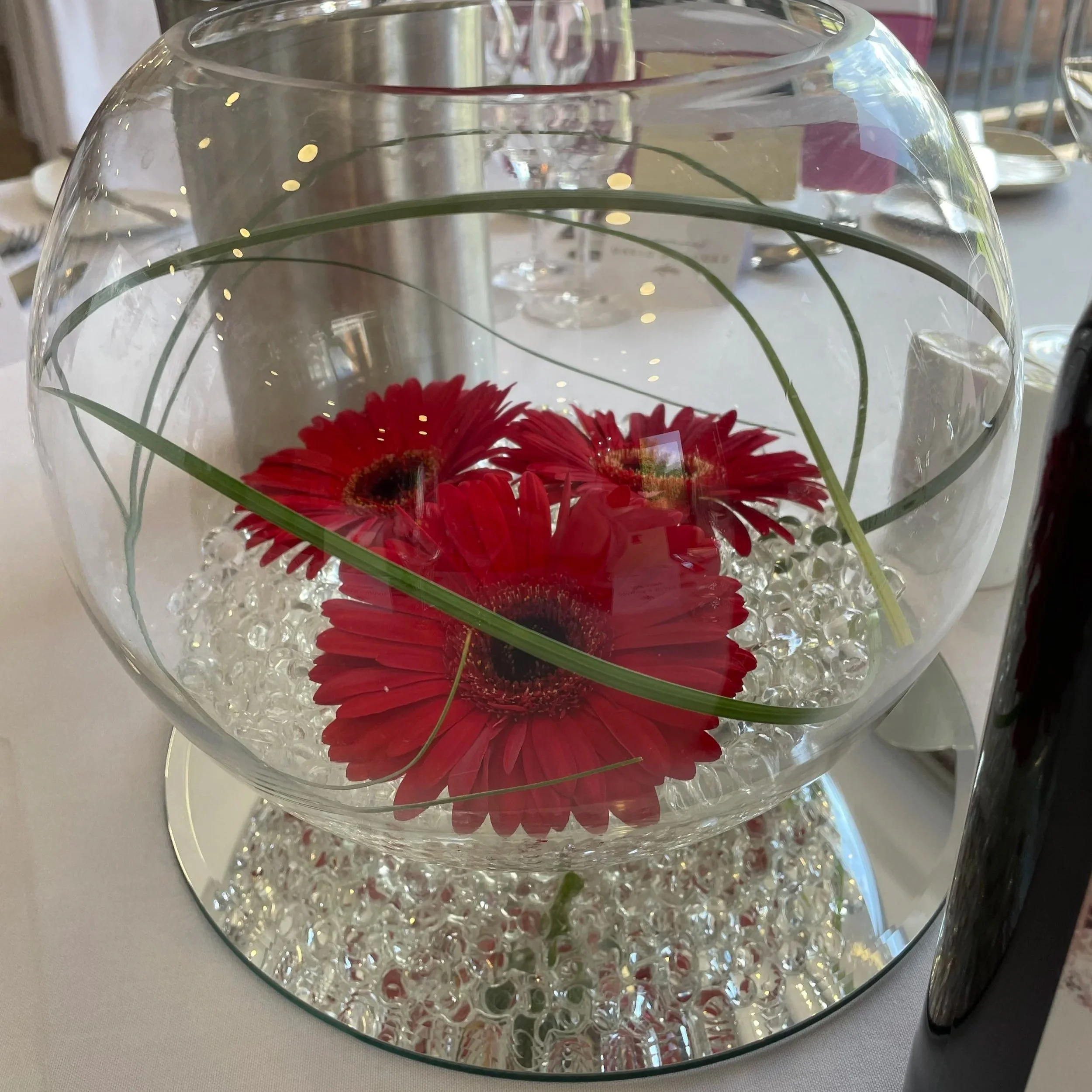 A glass bowl with red gerbera daisies and green grass inside, resting on a mirror base filled with clear decorative beads.