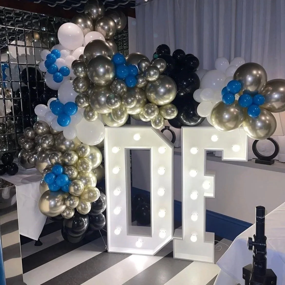 Large illuminated numbers '01' surrounded by a balloon arch with black, white, gold, and blue balloons at a celebration or event.
