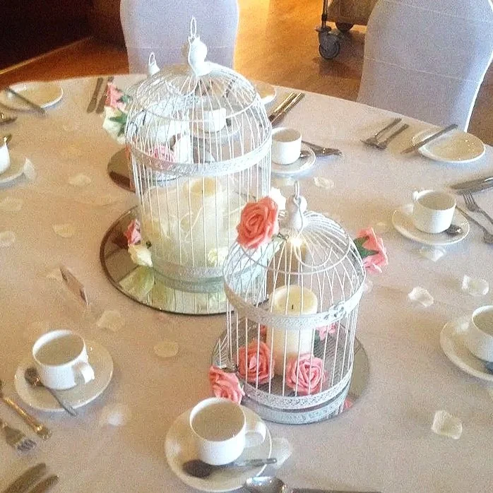 Decorative table setting with two white birdcages filled with pink and white roses, surrounded by white cups and saucers, teaspoons, and scattered flower petals.
