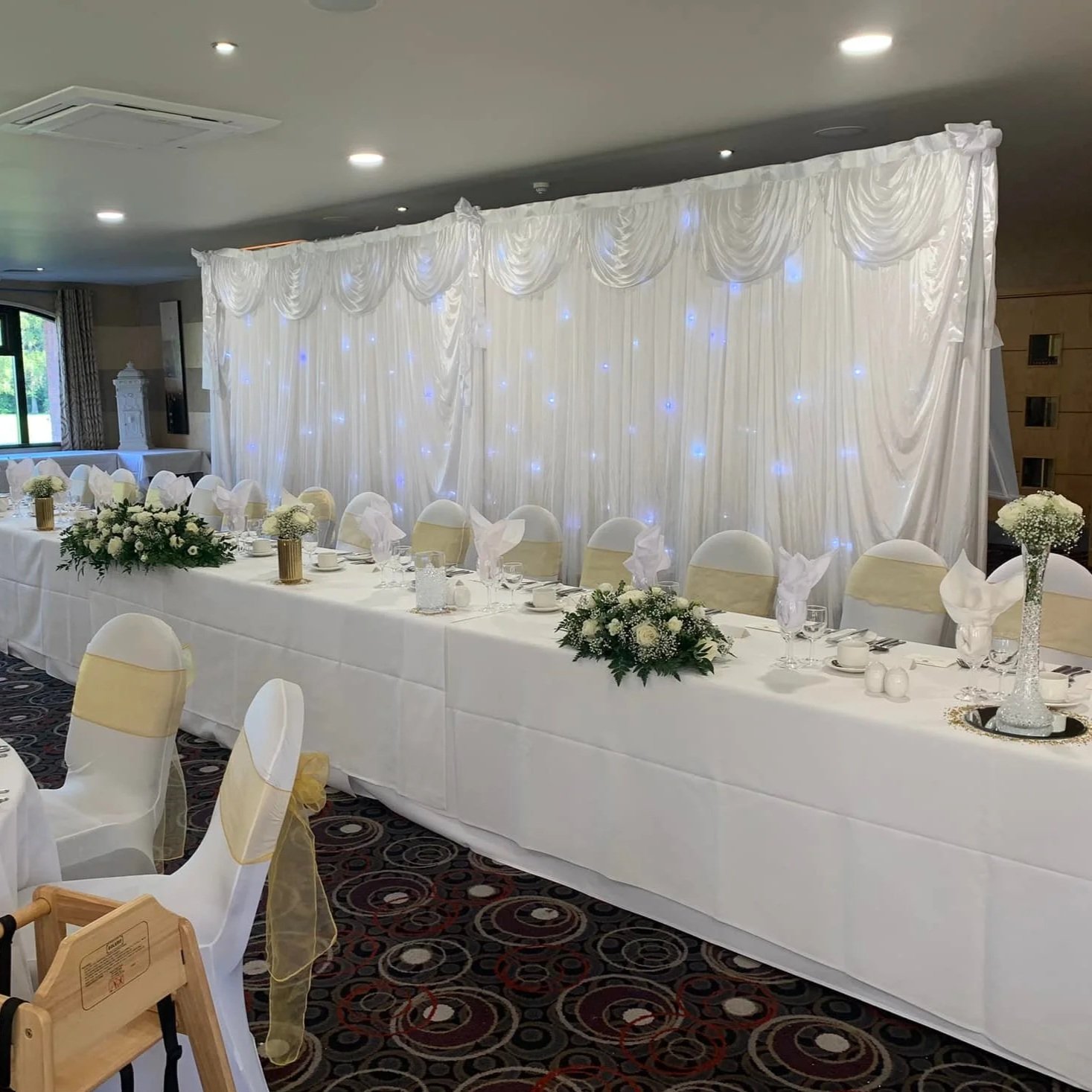 Elegant wedding reception setup with a long white bridal table, white chairs with gold sashes, floral centerpieces, and a white fabric backdrop with blue lights.