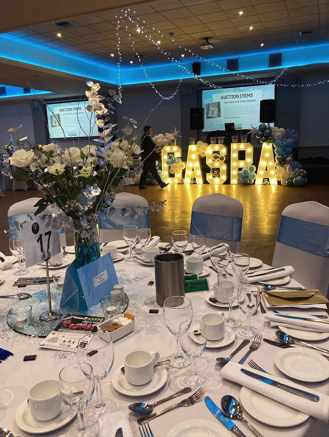 A banquet hall decorated with a large illuminated sign spelling 'GASPA' and filled with balloons. There is a stage in the background with a presentation slide that reads 'Auction Items' and a woman walking nearby. The foreground shows a round table s