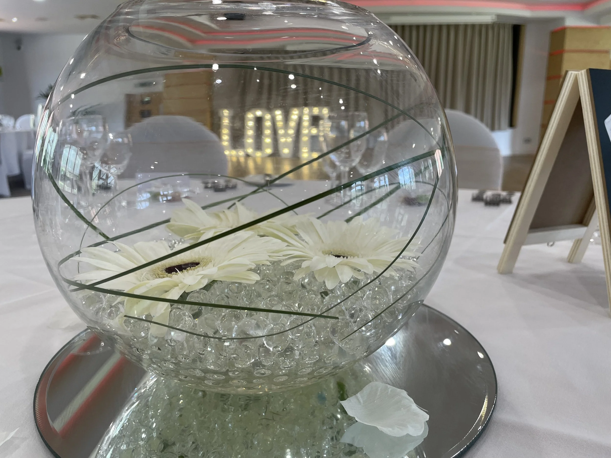 Glass bowl centerpiece with white flowers and glass beads on a table at an event or wedding reception.