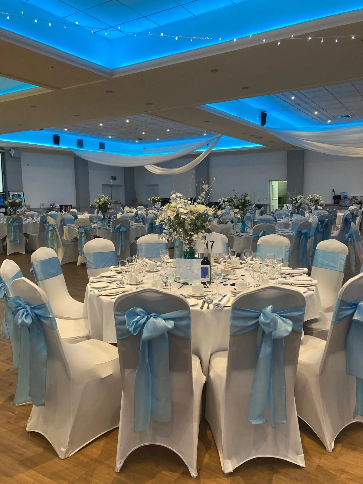 Elegant banquet hall decorated with white and blue theme, round tables with white tablecloths, white chairs adorned with blue bows, floral centerpieces, and blue ceiling lighting.