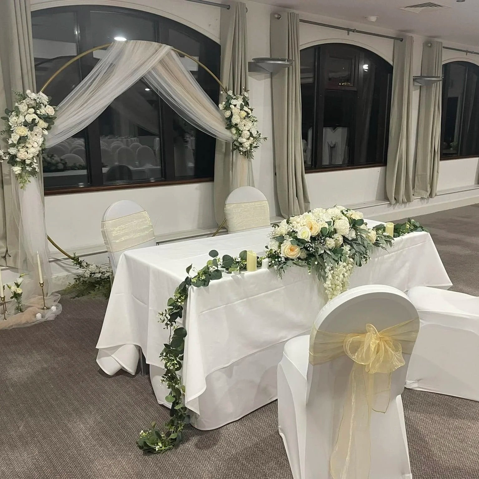 Wedding reception table decorated with white flowers, green foliage, candles, and gold accents, with a floral and fabric arch in the background.