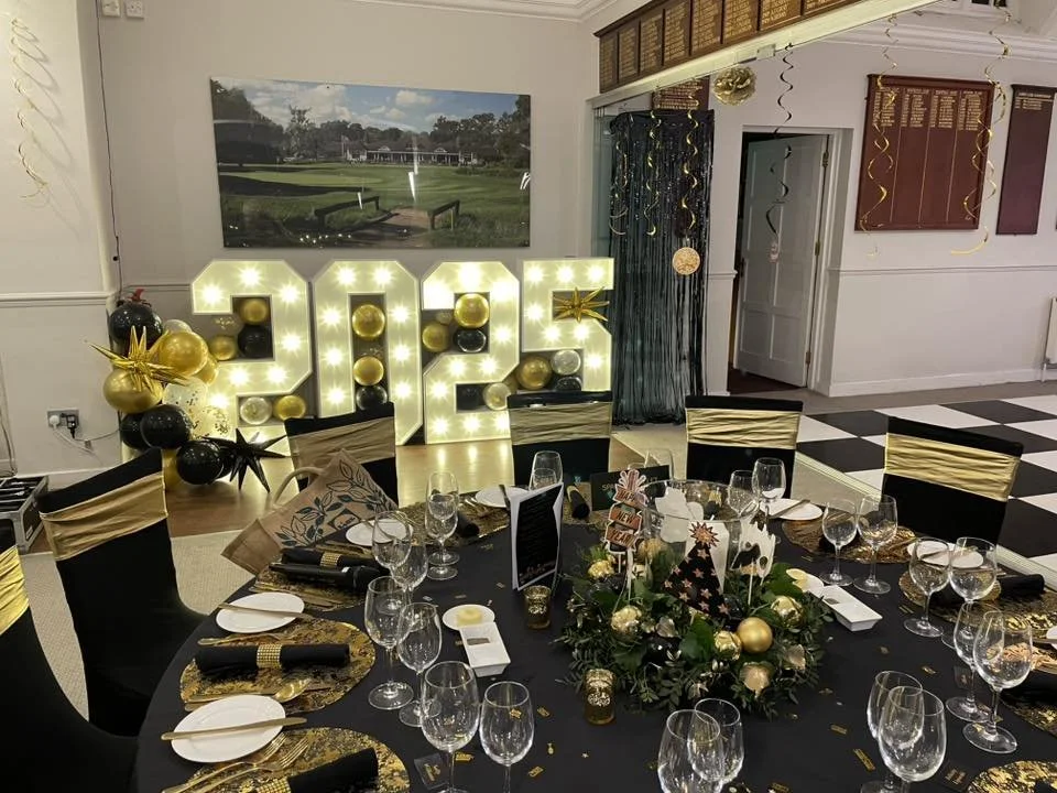 Party room decorated for New Year's celebration with a table set for guests, gold, black, and white color theme, large illuminated '2023' sign, decorated balloons, and festive decorations.
