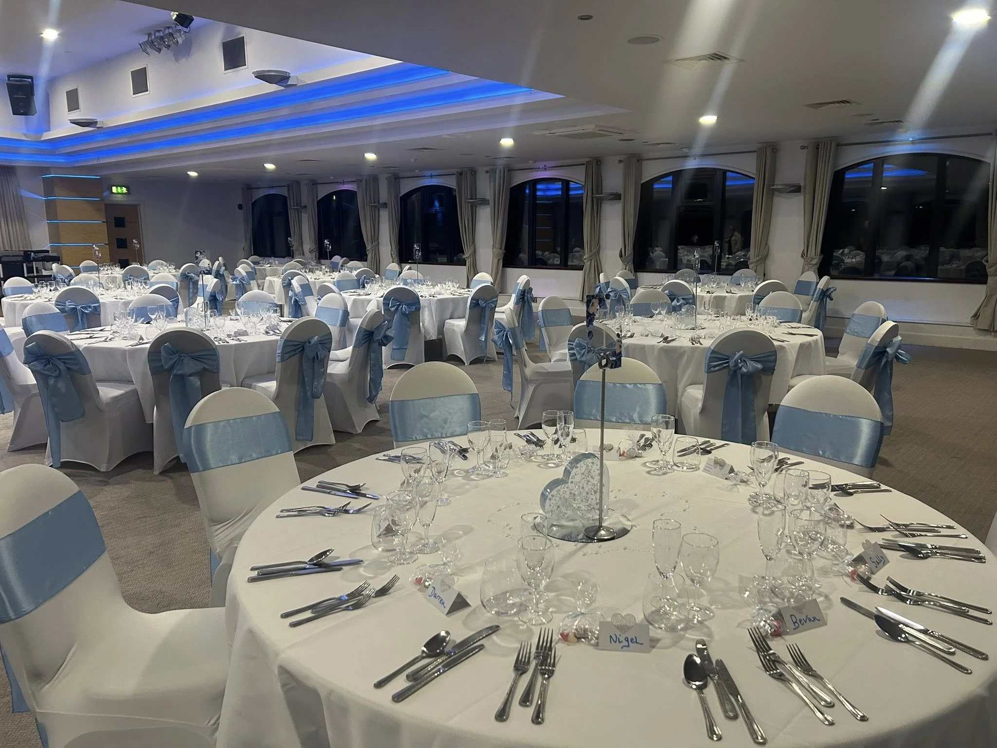 Banquet hall decorated for a formal event with round tables covered with white tablecloths and chairs draped in white with blue sashes. The tables are set with glassware, cutlery, and place cards. Blue lighting accents the ceiling, and large windows 