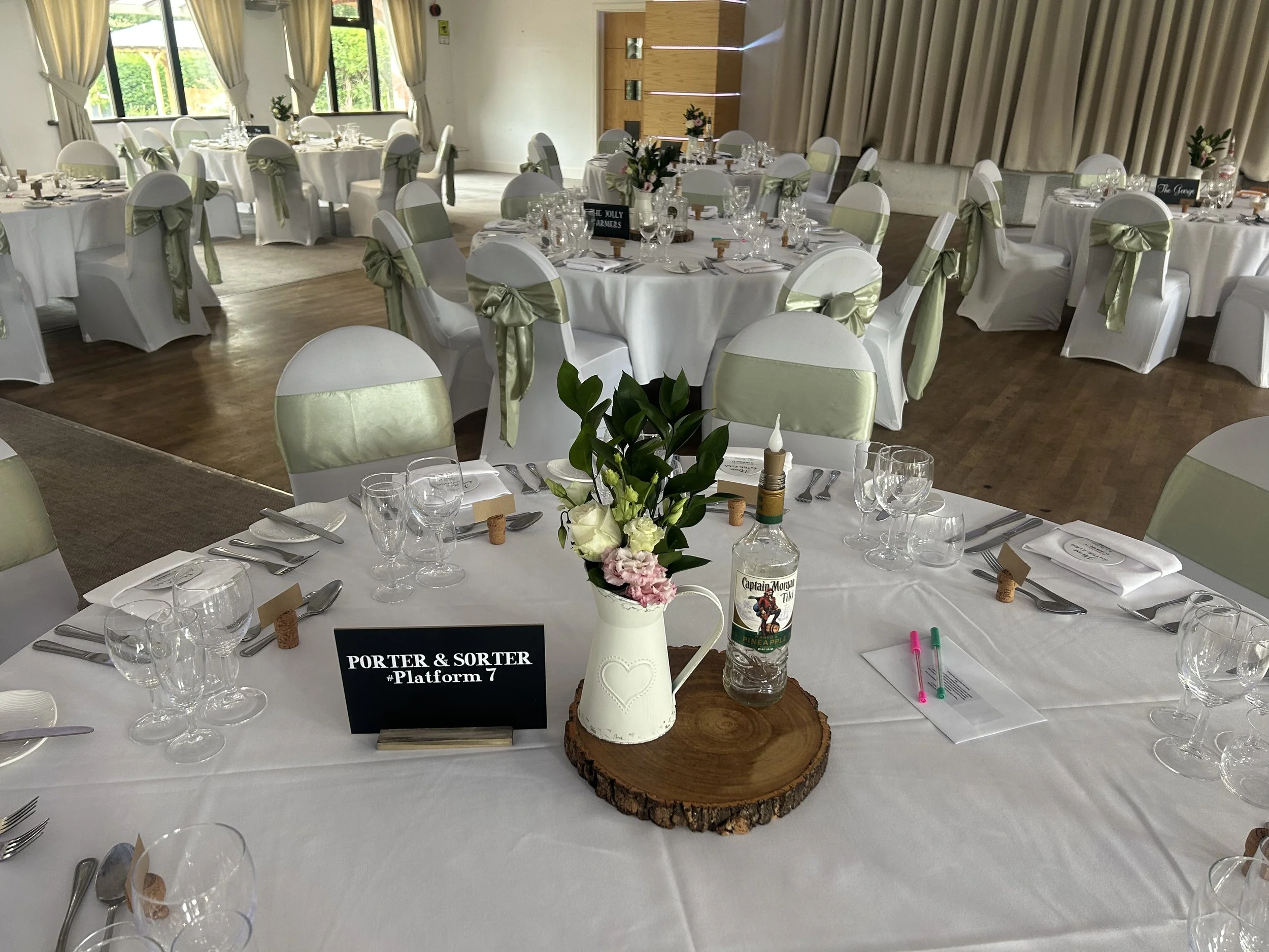 Wedding reception setup with round tables, white tablecloths, white chairs with green sashes, floral centerpieces, and a wooden slab centerpiece with a vase, bottle, and pink and white flowers.