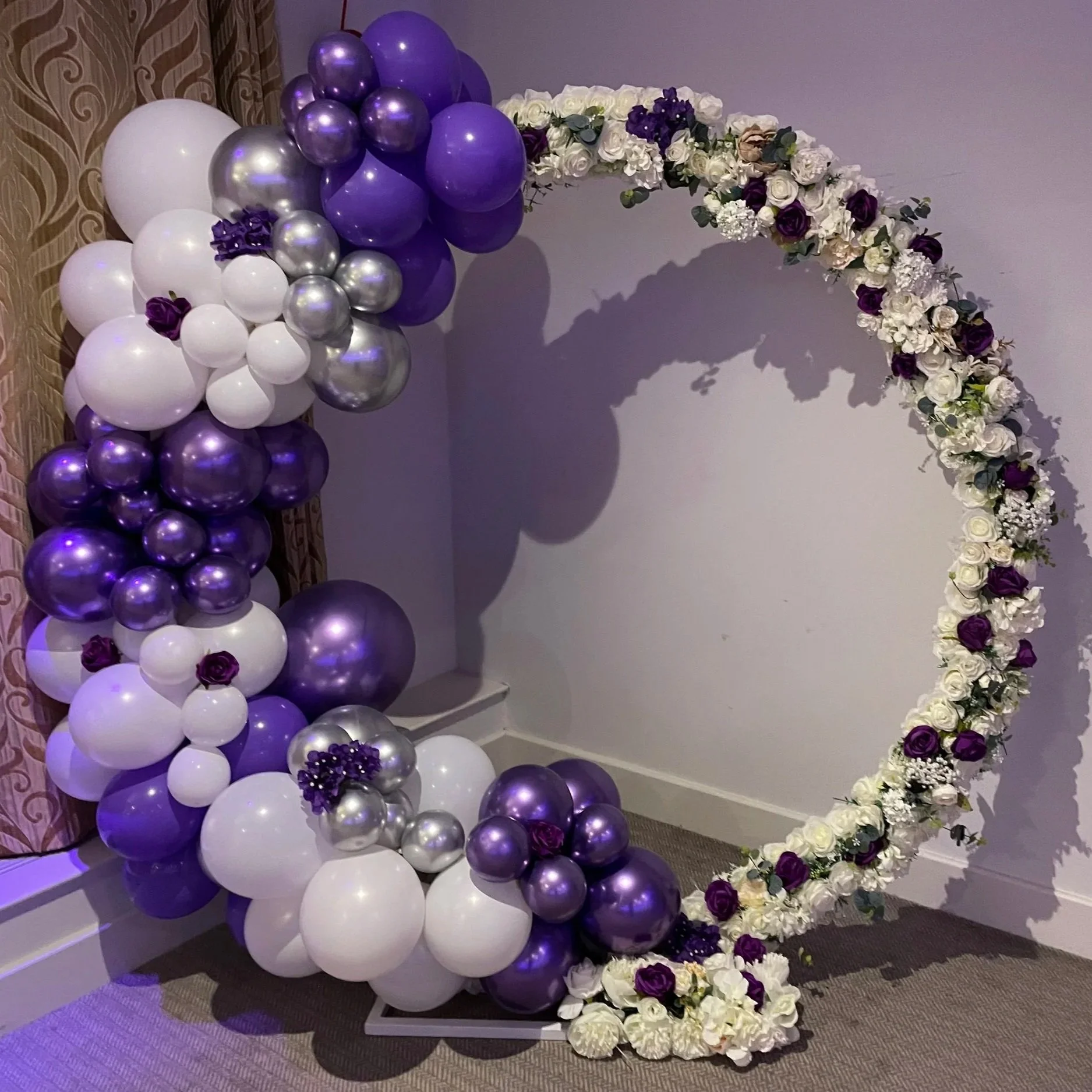 Decorative balloon arch with purple, white, and metallic balloons and flowers at the base, suitable for events like weddings or celebrations.