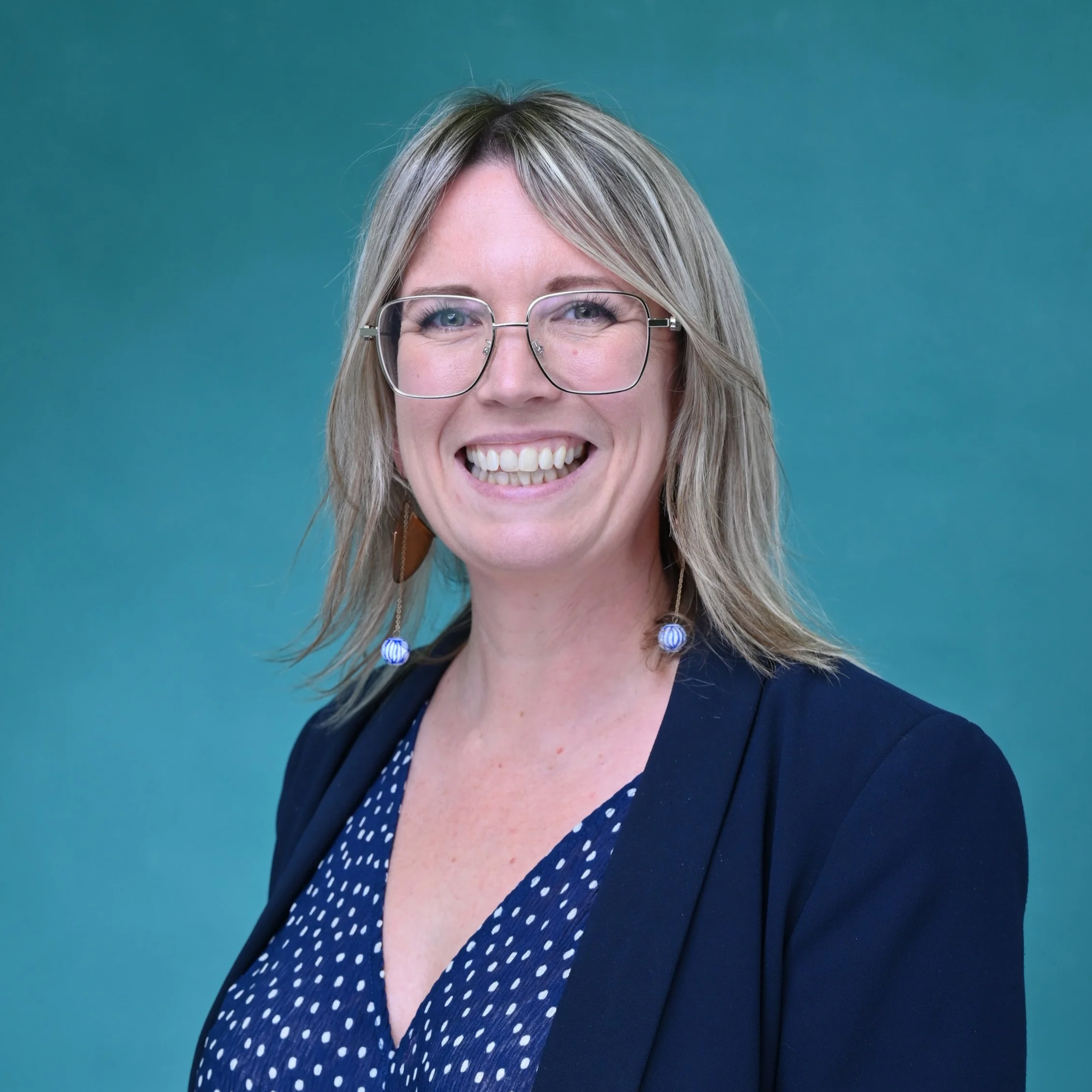 Lucy Lee Allen is a white woman with blonde hair. She is wearing  glasses and a blue polka dot dress nd navy blazer/ She is in front of a blue background