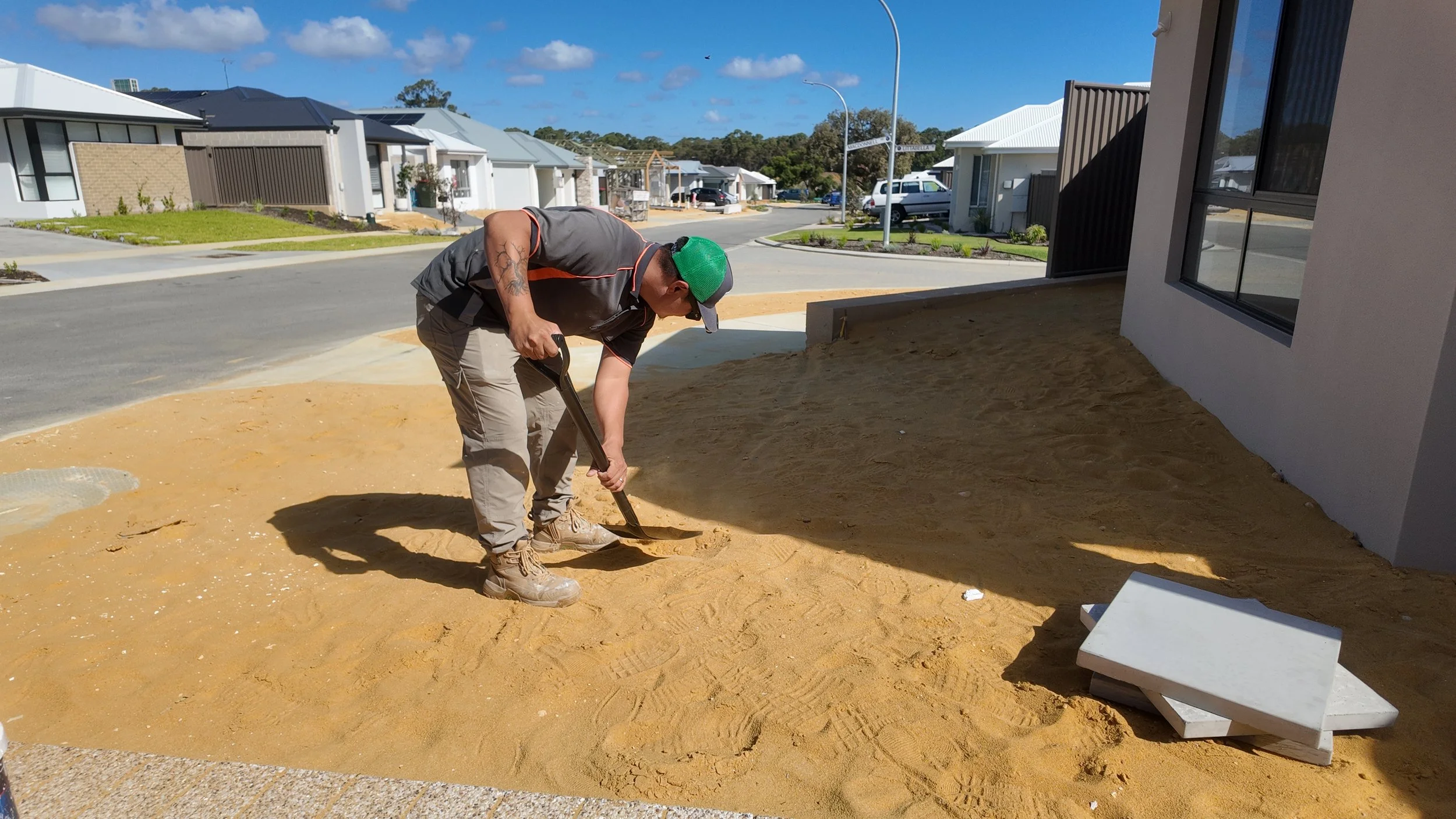 A man wearing a gray t-shirt, khaki pants, and a green baseball cap is digging in a yard with a shovel outside a modern house, with a residential street in the background under a blue sky.
