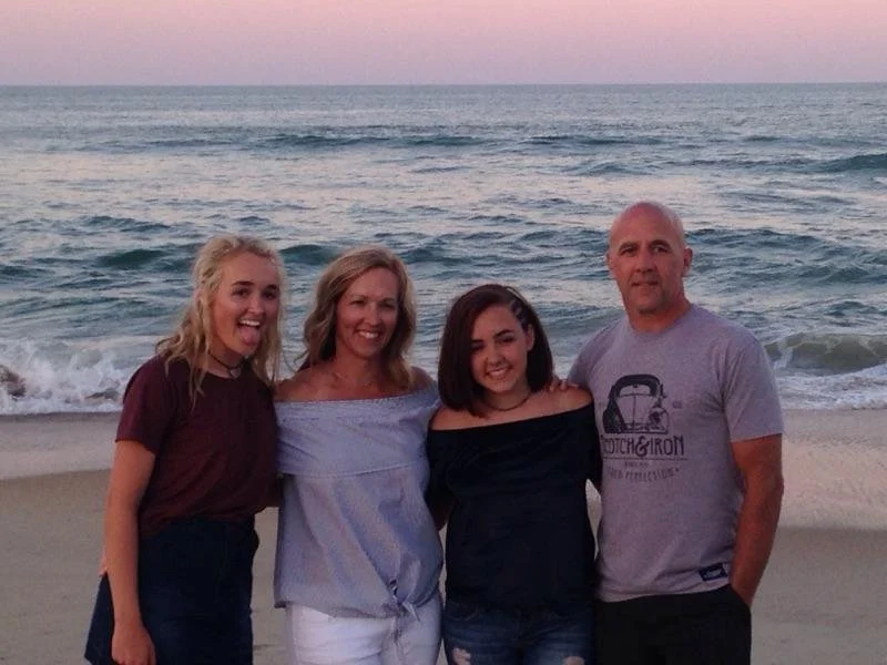 Family of four posing on the beach at sunset, with the ocean and pink sky in the background.
