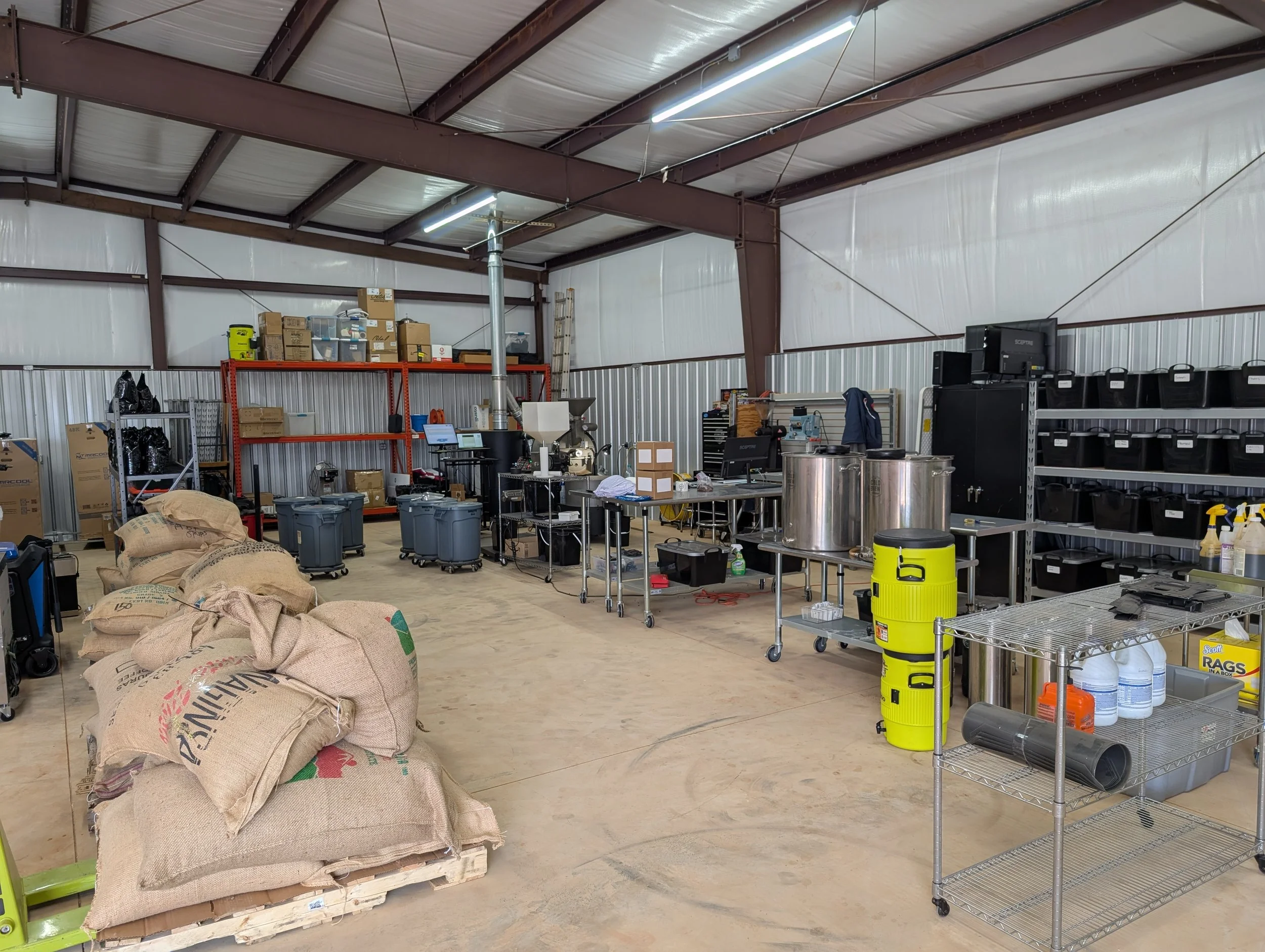 Inside a warehouse with metal walls and ceiling, containing shelves with boxes, bins, and equipment, a pallet with sacks, and cleaning supplies.