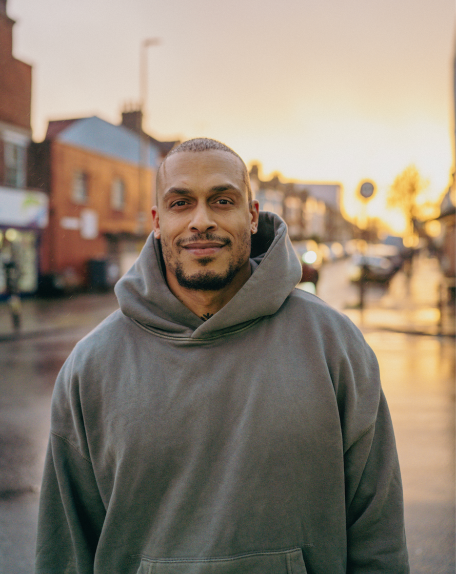 A man with a goatee wearing a gray hoodie standing outdoors during sunset or sunrise on a wet street in a town or city.