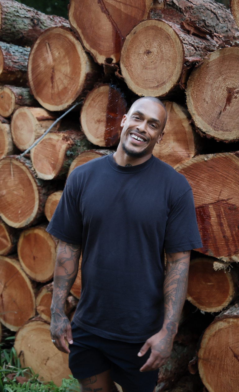 A smiling man with tattoos wearing a black t-shirt and shorts standing in front of a large pile of cut logs.