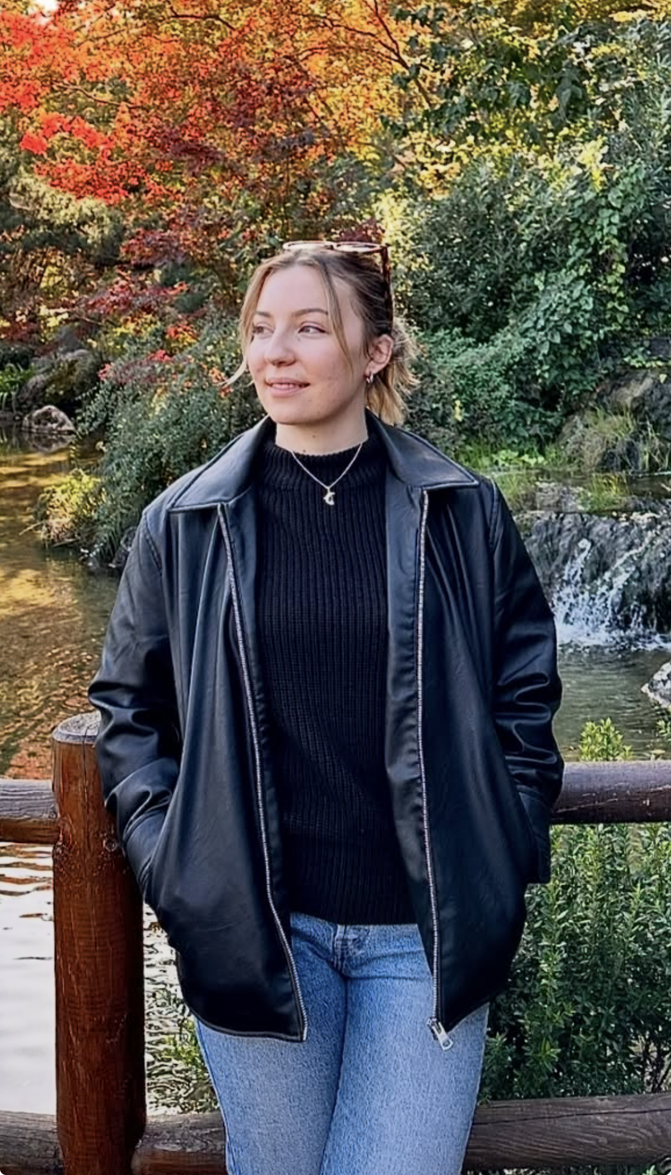A woman standing outdoors by a wooden railing, with colorful fall foliage in the background. She is wearing a black leather jacket, a black ribbed sweater, blue jeans, and a necklace, with sunglasses resting on her head.