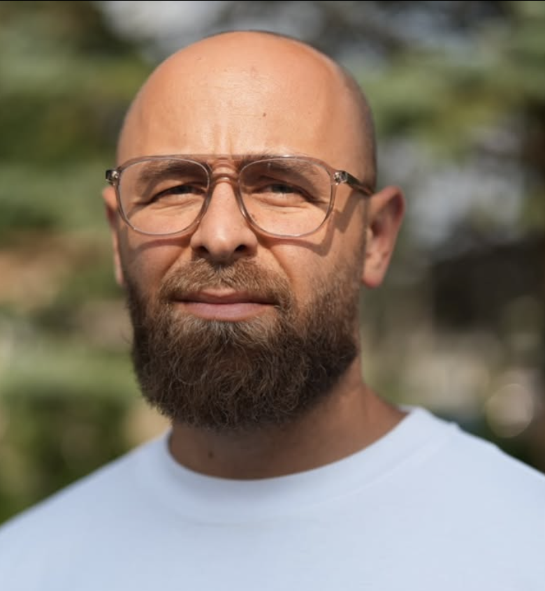 A middle-aged man with a beard wearing glasses and a white shirt outdoors with a blurred green background.